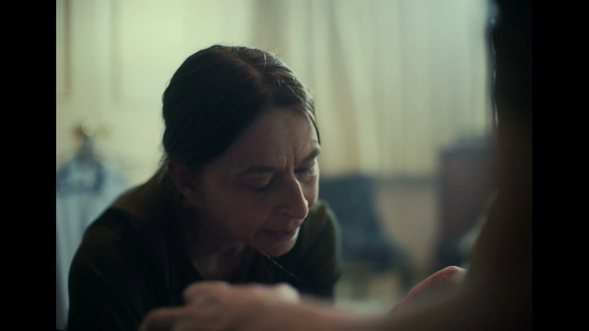 A woman with dark hair and a dark shirt looks distressed and confused, speaking to someone off-screen in a dimly lit room.