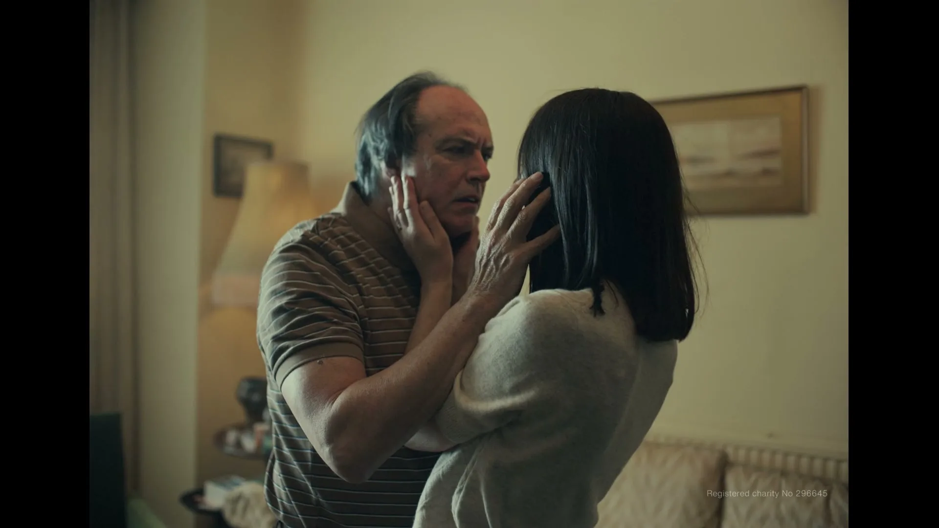 A woman with dark hair holds the face of an older man with a concerned expression in a dimly lit living room.