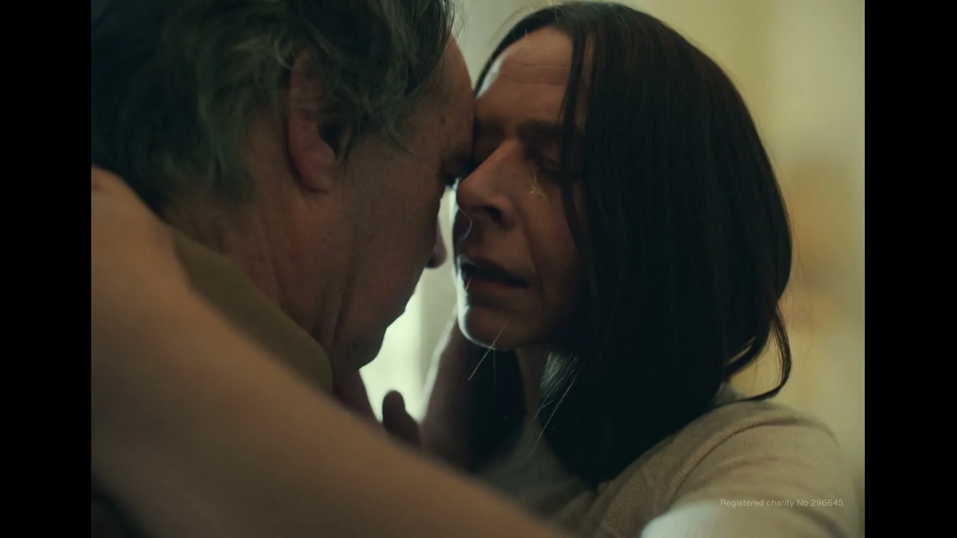 A middle-aged man and woman embrace and kiss, the woman crying, in a softly lit interior.