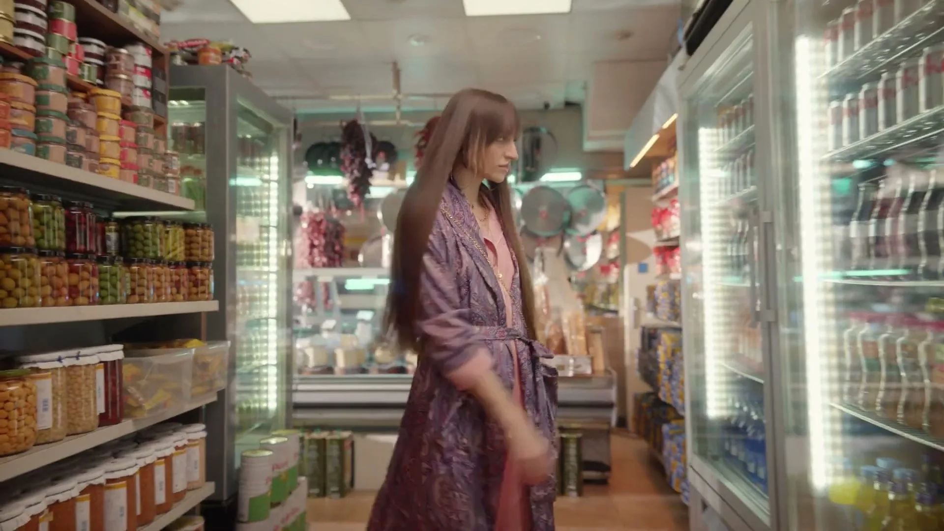A young woman with long brown hair, wearing a patterned robe over pink pajamas, walks through a small grocery store aisle and opens a refrigerator door.