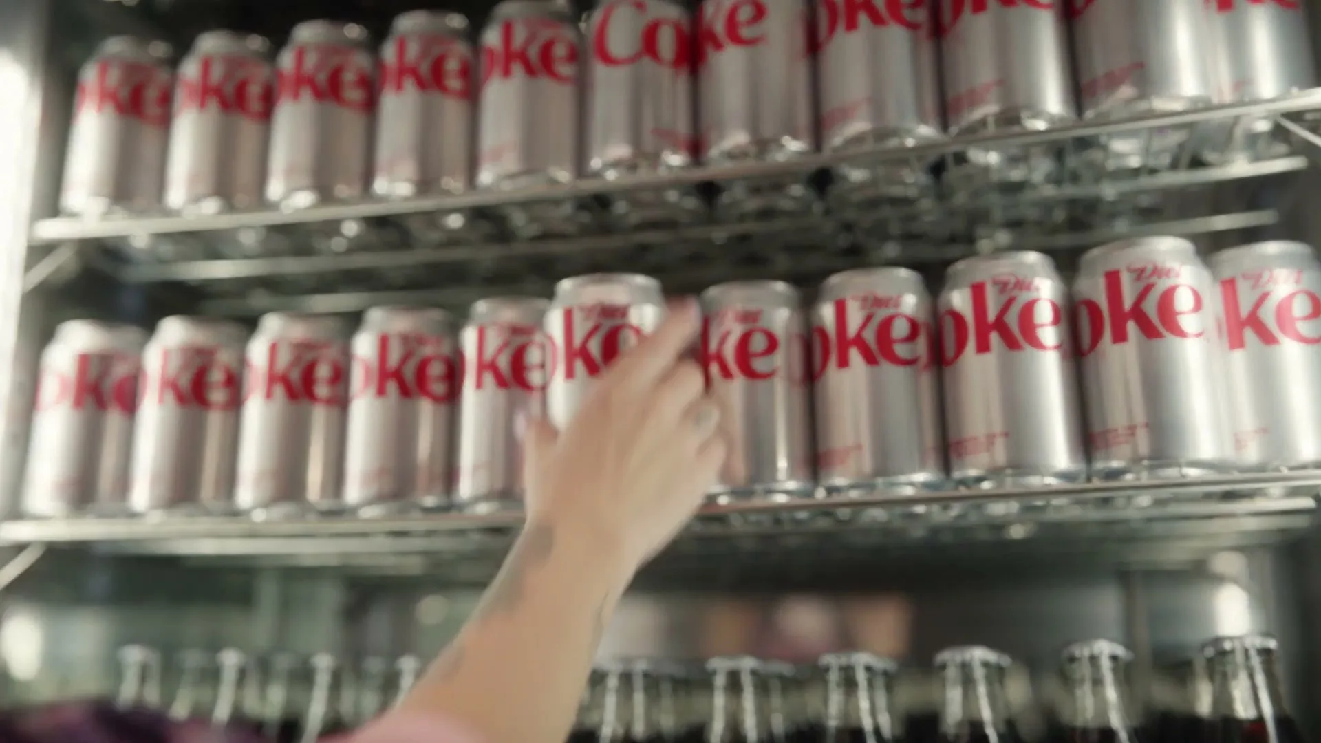 A person's hand reaches into a refrigerated display case filled with Diet Coke cans and bottles, selecting a can.