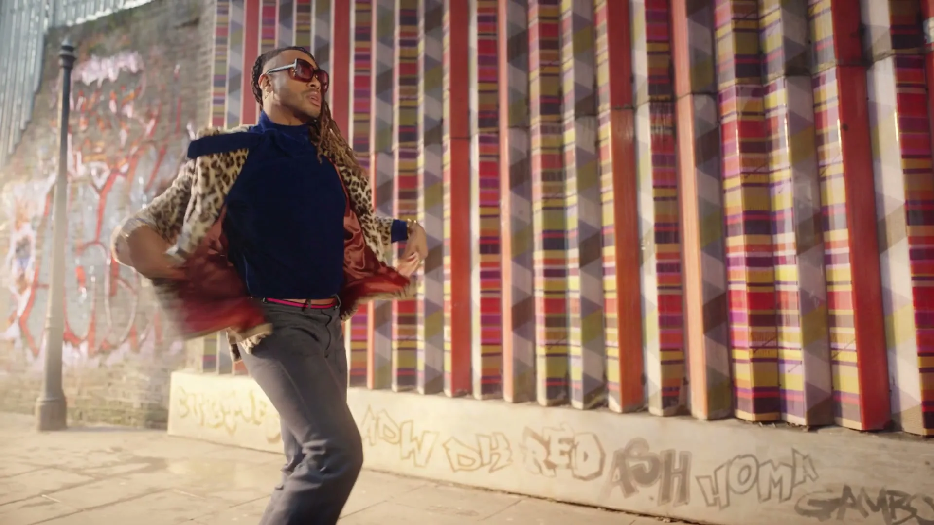 A man with dreadlocks, wearing a leopard print coat and sunglasses, dances energetically on a city street next to a colorful striped wall.