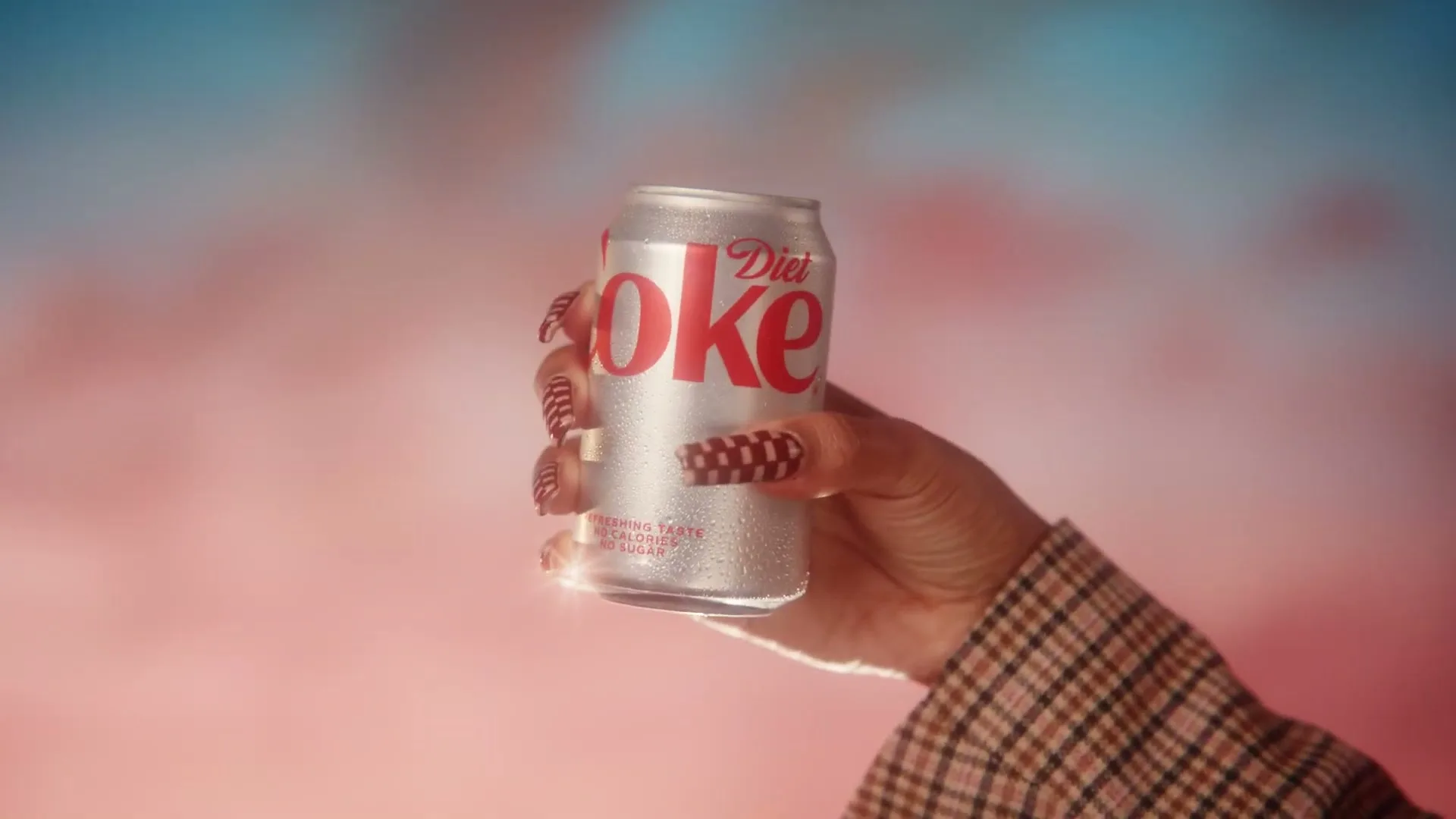 A hand with checkered nails holds a cold Diet Coke can against a soft-focus background of pink and blue clouds.