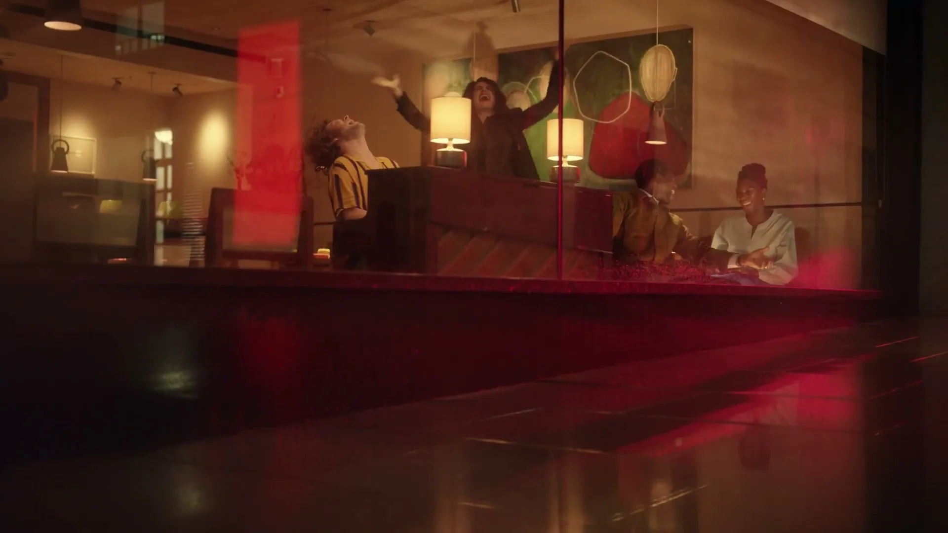 Four friends are seen through a window, laughing and dancing around a piano in a warmly lit room.