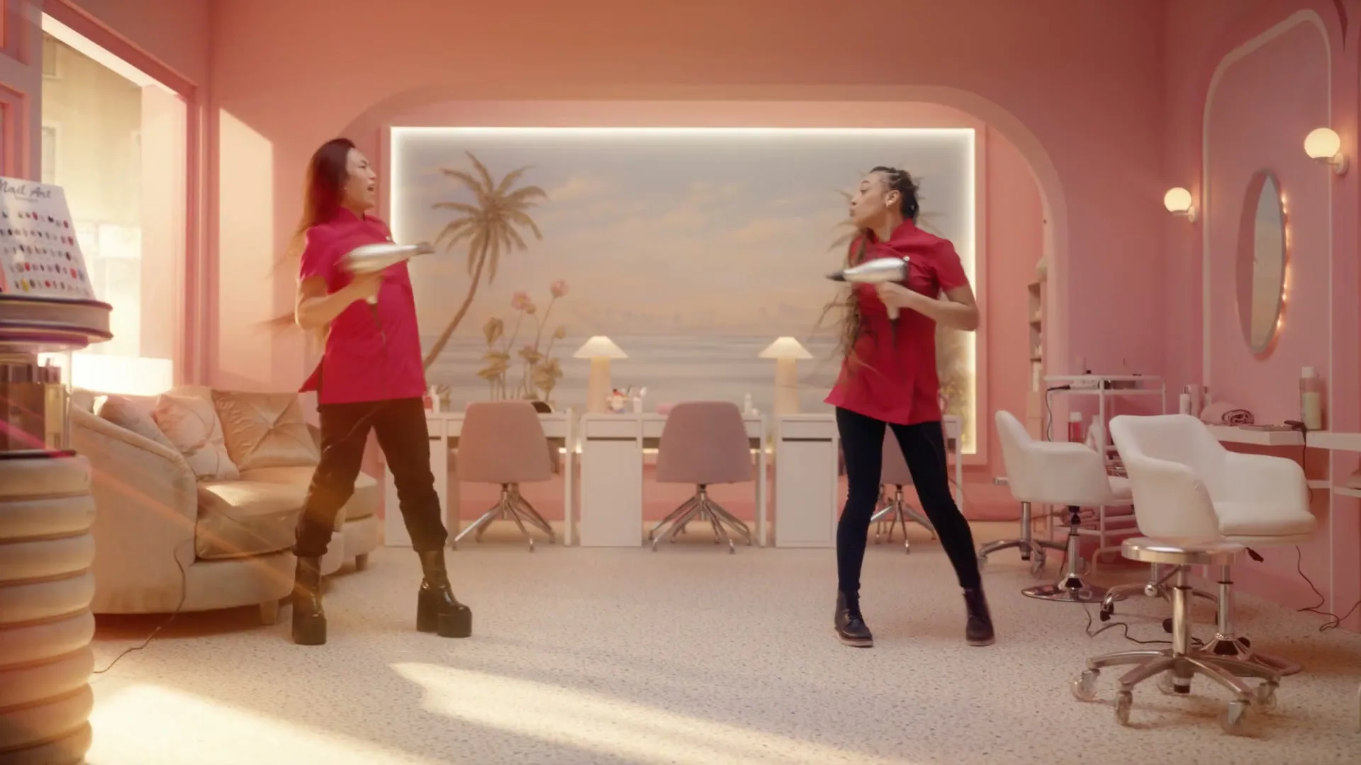 Two women in red shirts and dark pants dance playfully with hair dryers in a pink-themed nail salon.