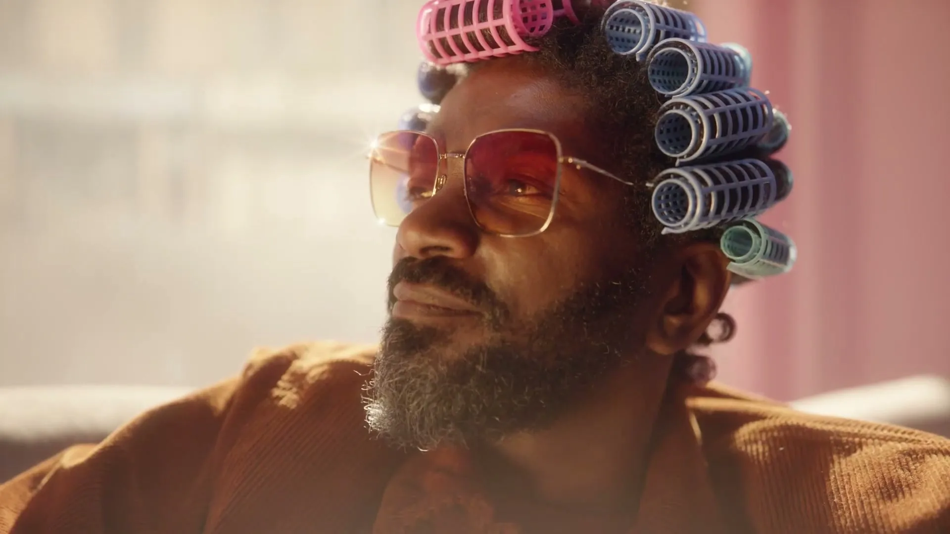 A man with a beard and sunglasses, wearing hair rollers, looks off-screen in a sunlit room.