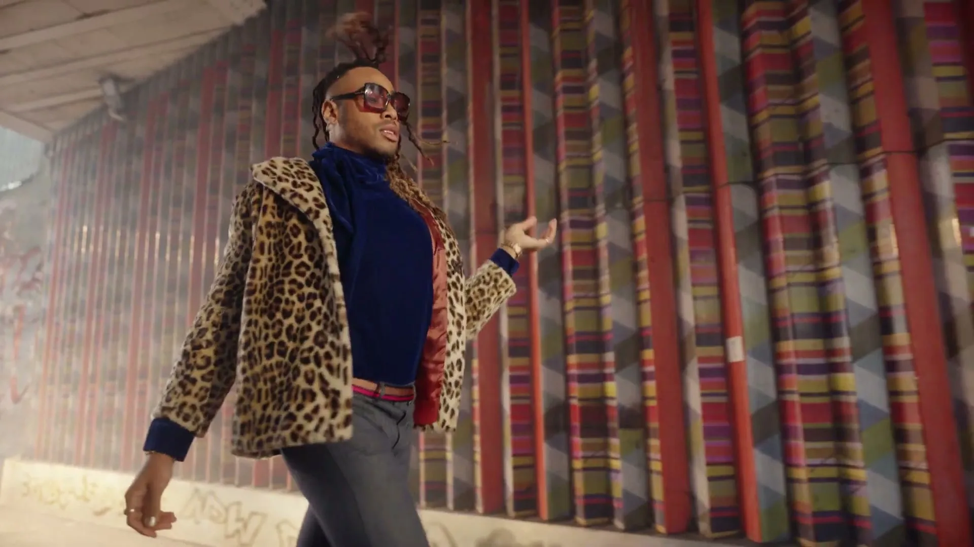 A person in a leopard print coat and sunglasses walks confidently past a colorful, striped wall in an urban underpass.