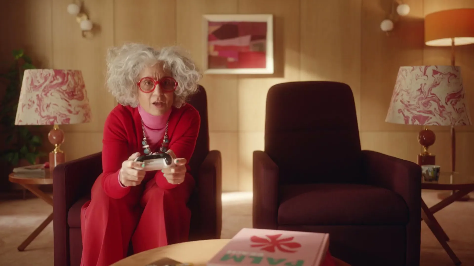 A senior woman with curly grey hair and red glasses, wearing a red outfit, sits in an armchair playing a video game in a retro living room.