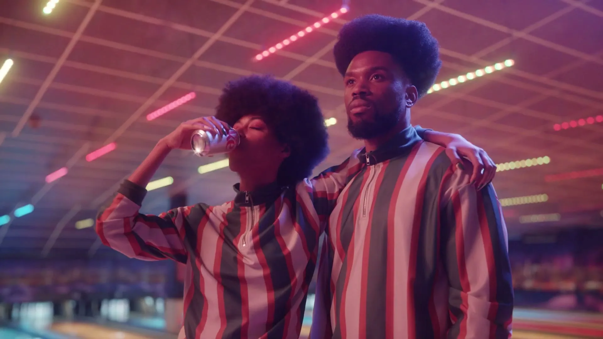 A young Black woman with an afro drinks from a Coca-Cola can, standing next to a young Black man with an afro, both wearing matching striped shirts in a neon-lit bowling alley.