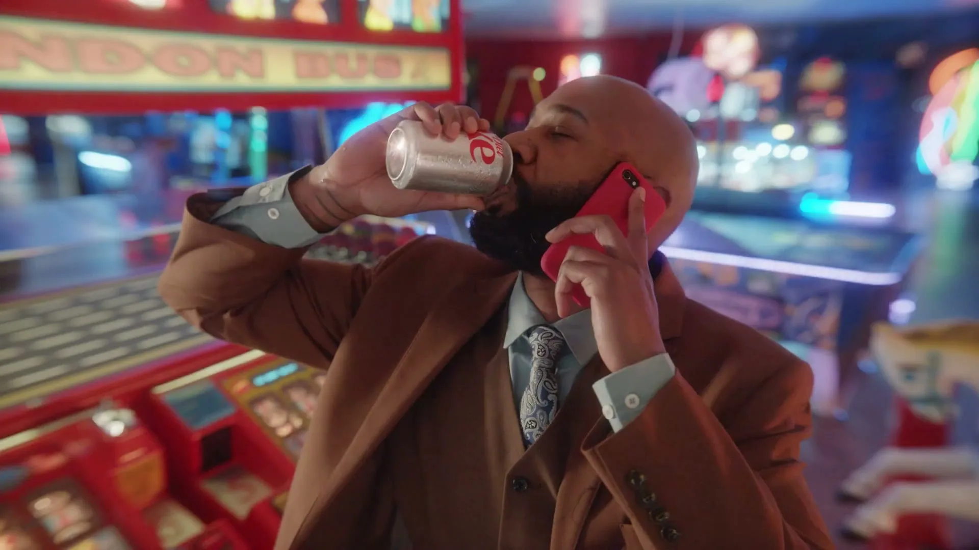 A bald man with a beard, wearing a brown suit, drinks a Coca-Cola while talking on a red phone in a neon-lit arcade.