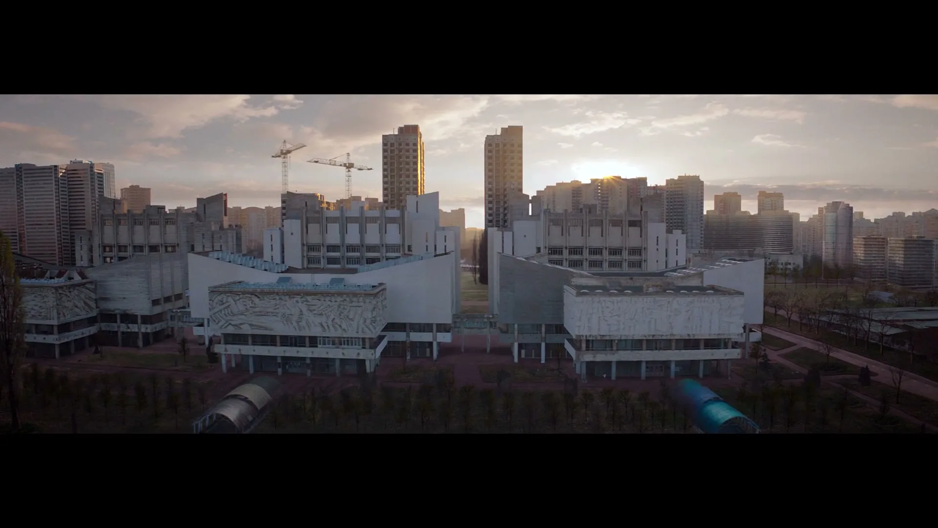 An aerial wide shot captures a dystopian city with brutalist architecture and construction cranes under a dramatic sky at sunset.