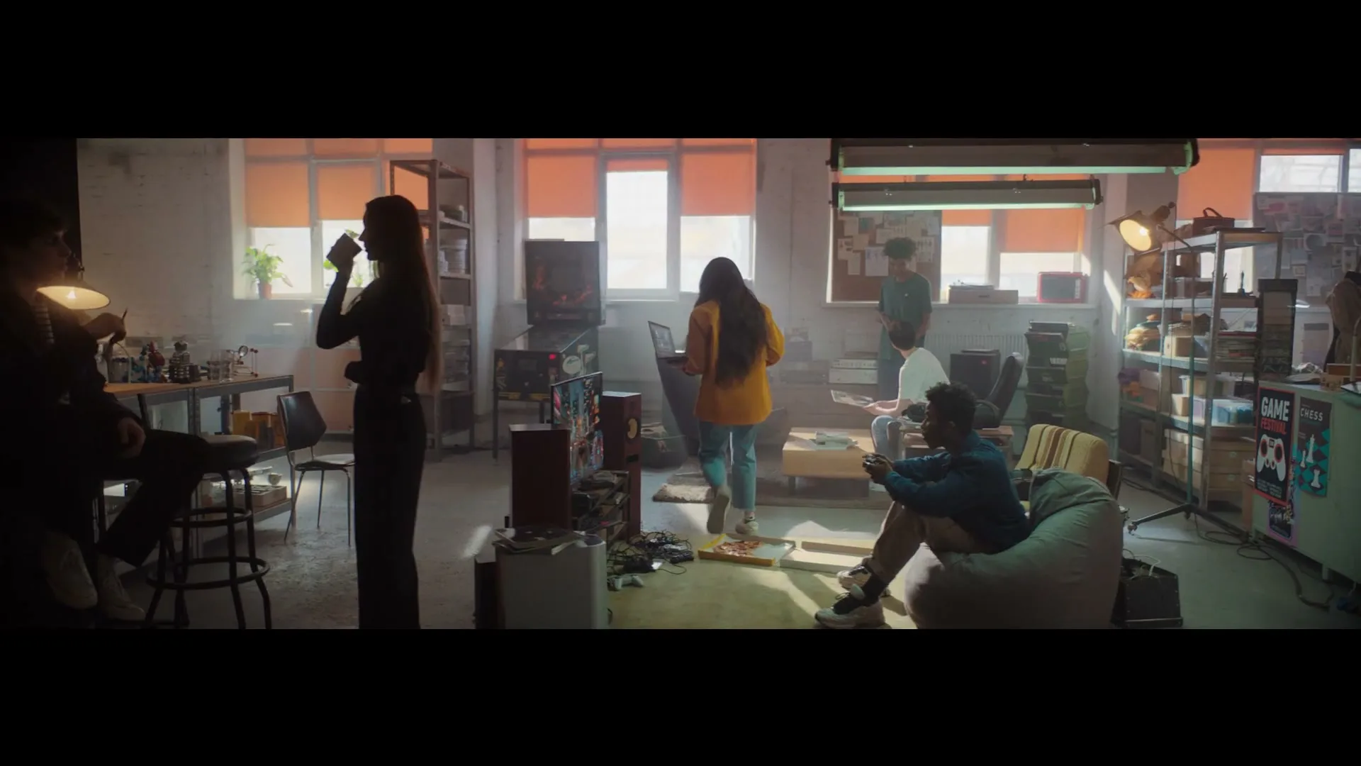 A diverse group of young adults are hanging out in a sunlit, industrial-style common room, some standing, some sitting, some looking at screens.