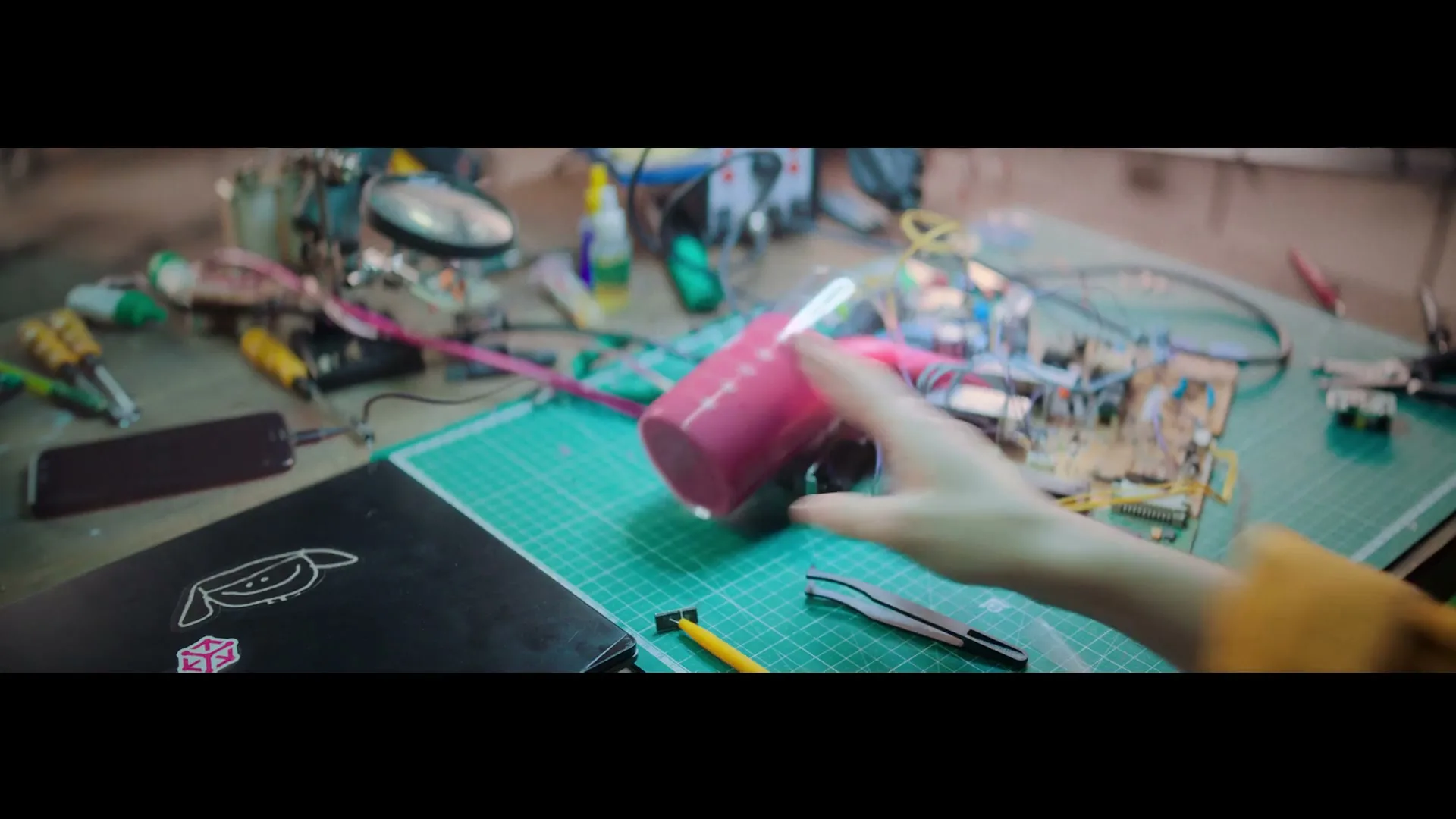A person's hand accidentally knocks over a pink bottle, causing sparks to fly from a circuit board on a cluttered workbench.