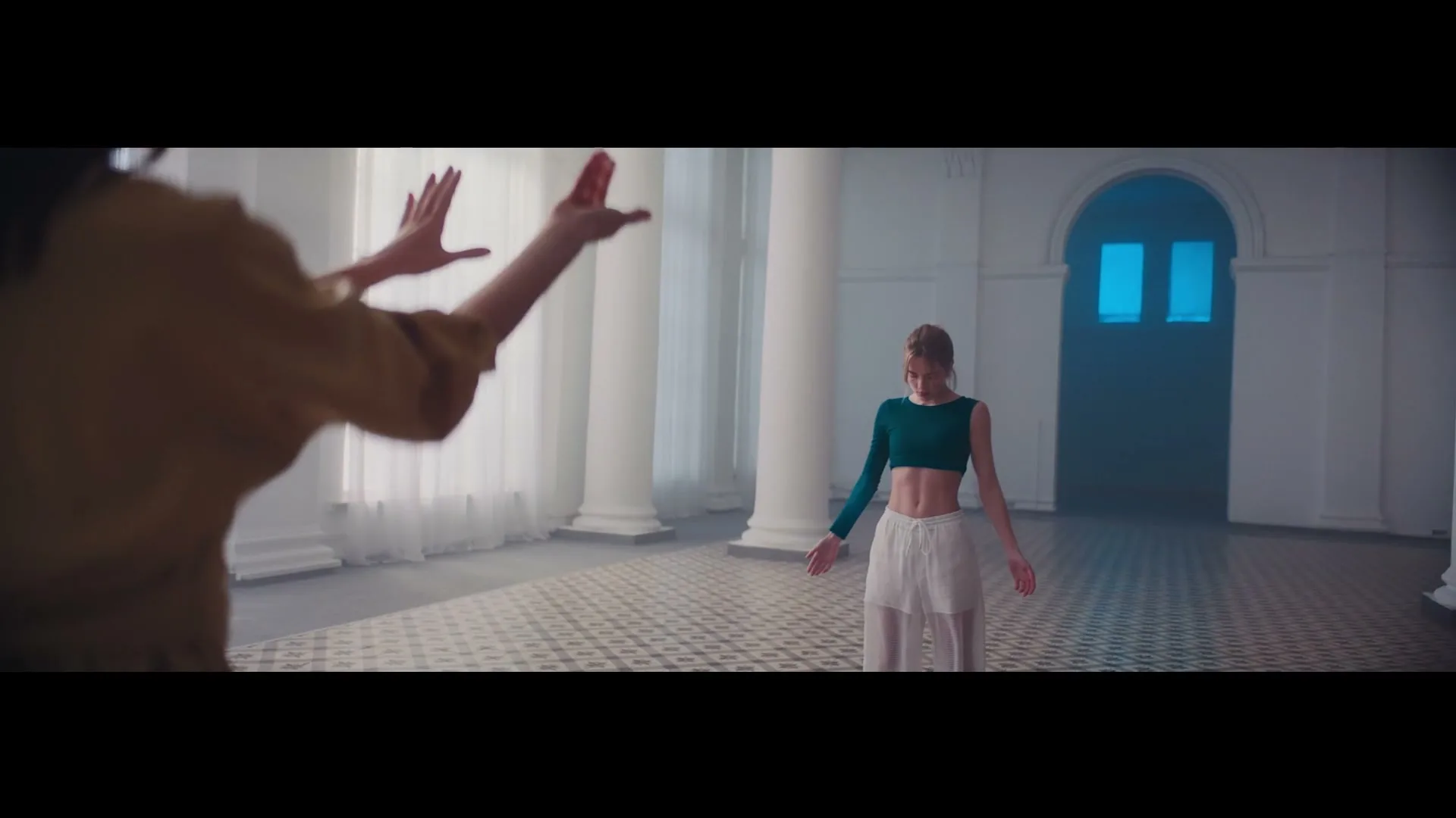 A young woman in a crop top and flowy pants dances in a large, white room with columns and an arched doorway, as another person's hands gesture in the foreground.