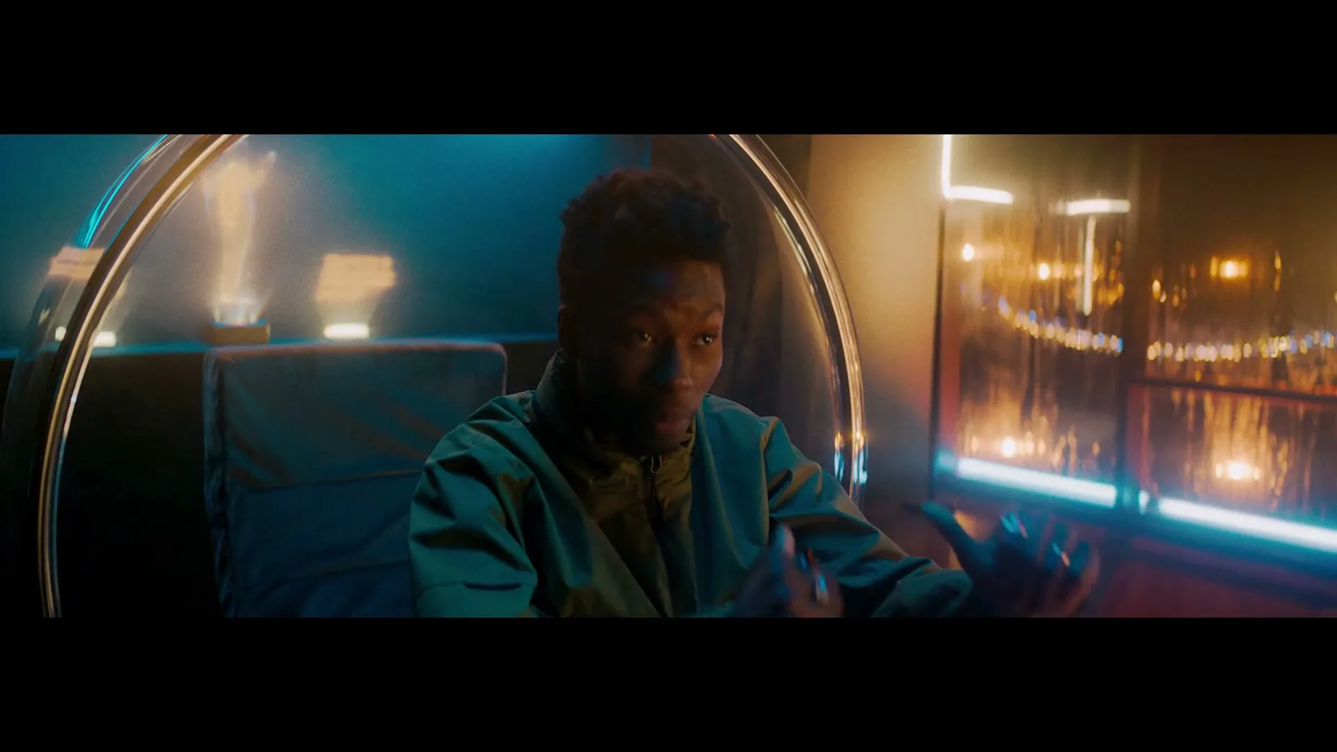 A young Black man with short dark hair, wearing a green jacket, sits in a futuristic chair, gesturing with his hands in a dimly lit, high-tech room.