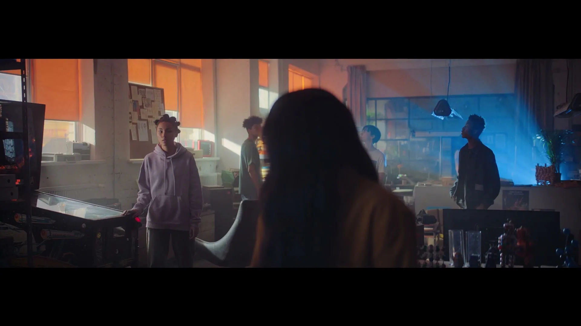 A diverse group of young adults stands in a sunlit, industrial-style room with a pinball machine, looking around with curiosity.