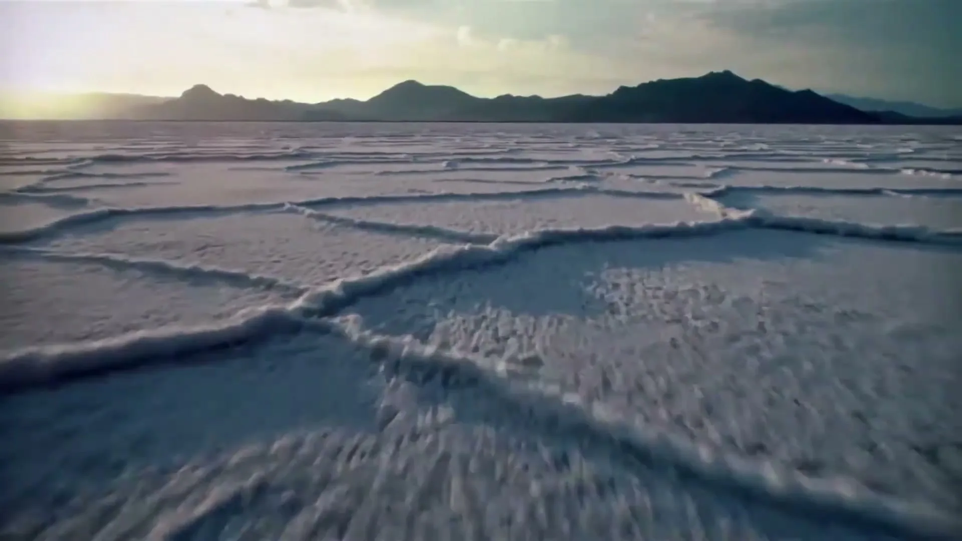 An aerial shot captures the vast, cracked surface of a salt flat, with distant mountains under a dramatic sky at golden hour.