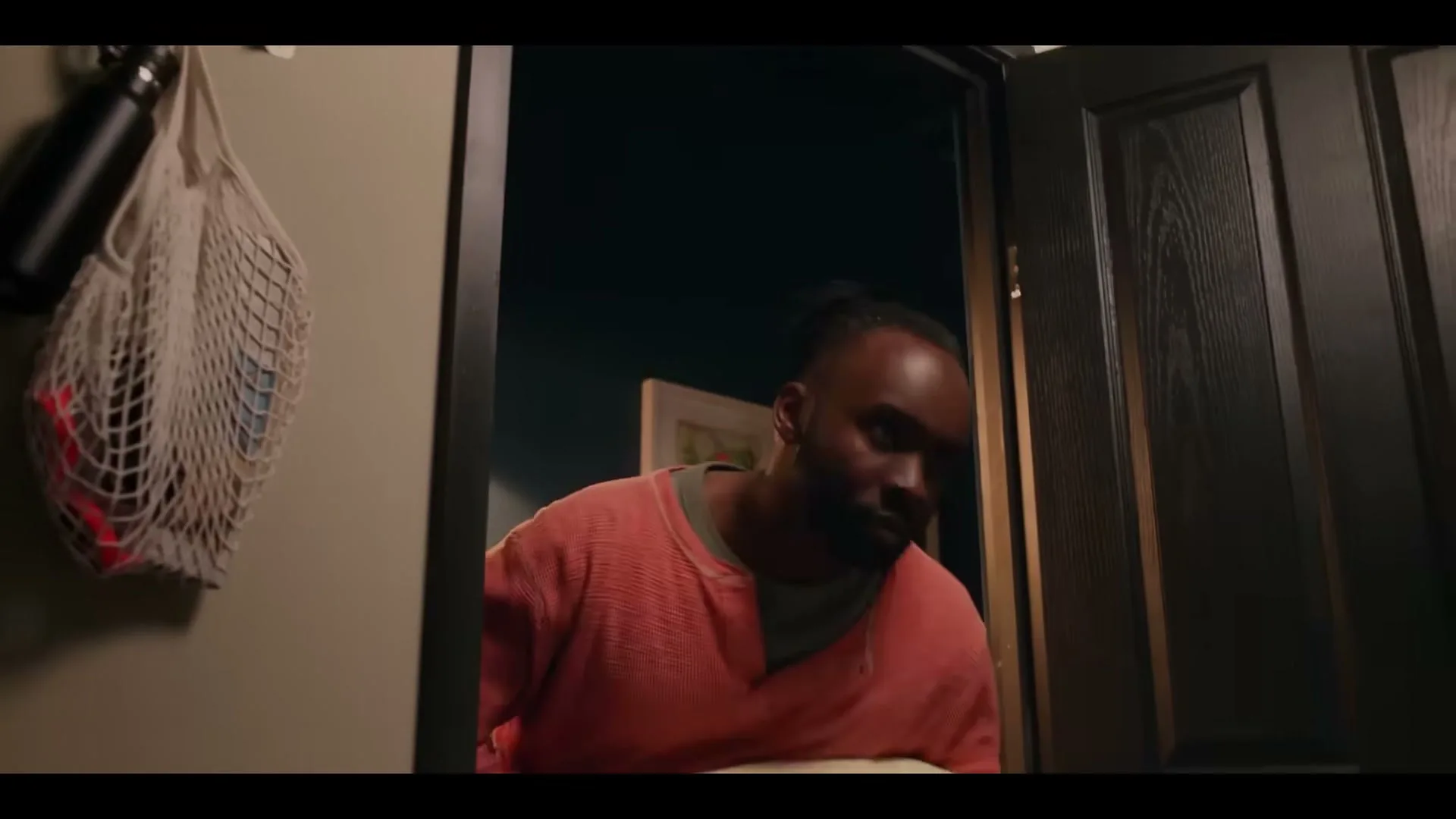 A man in a coral shirt enters a room through a dark door, carrying a large, light-colored pillow.