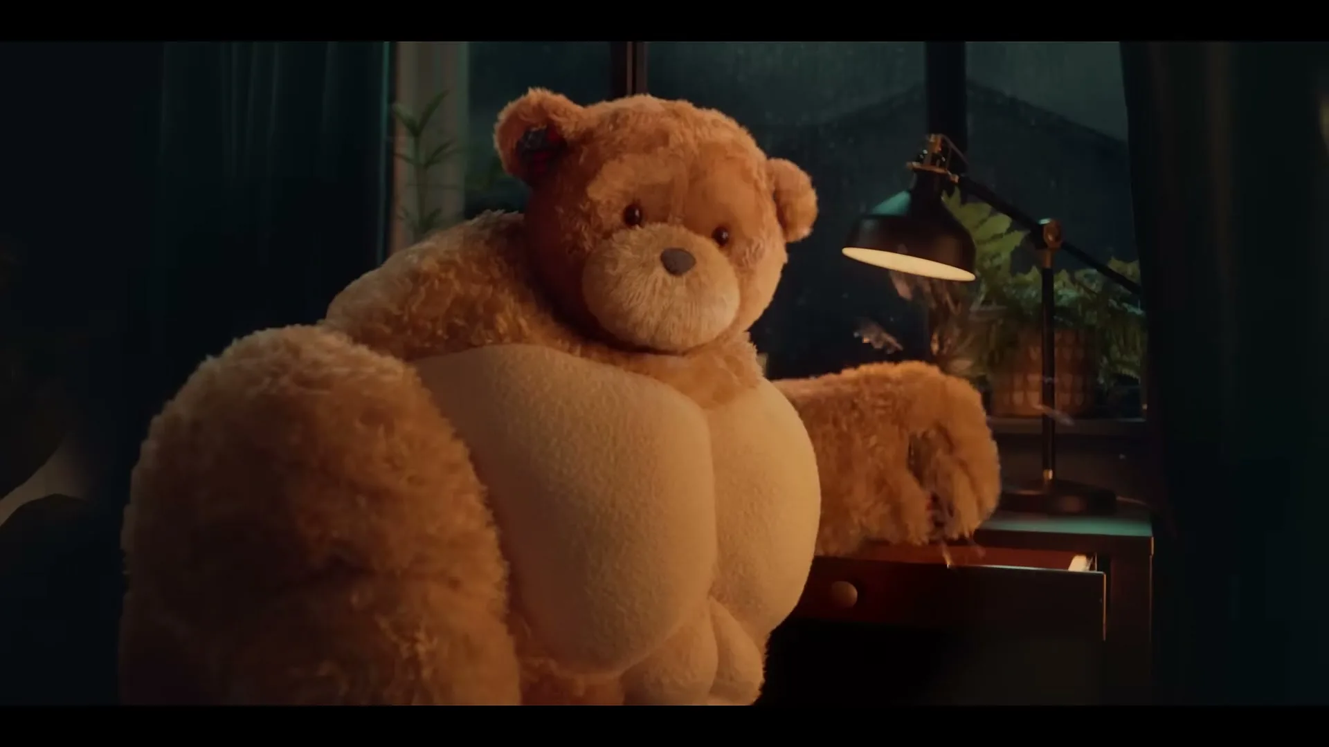 A large, muscular teddy bear sits at a desk, opening a drawer and looking at the camera in a dimly lit room during a rainy night.