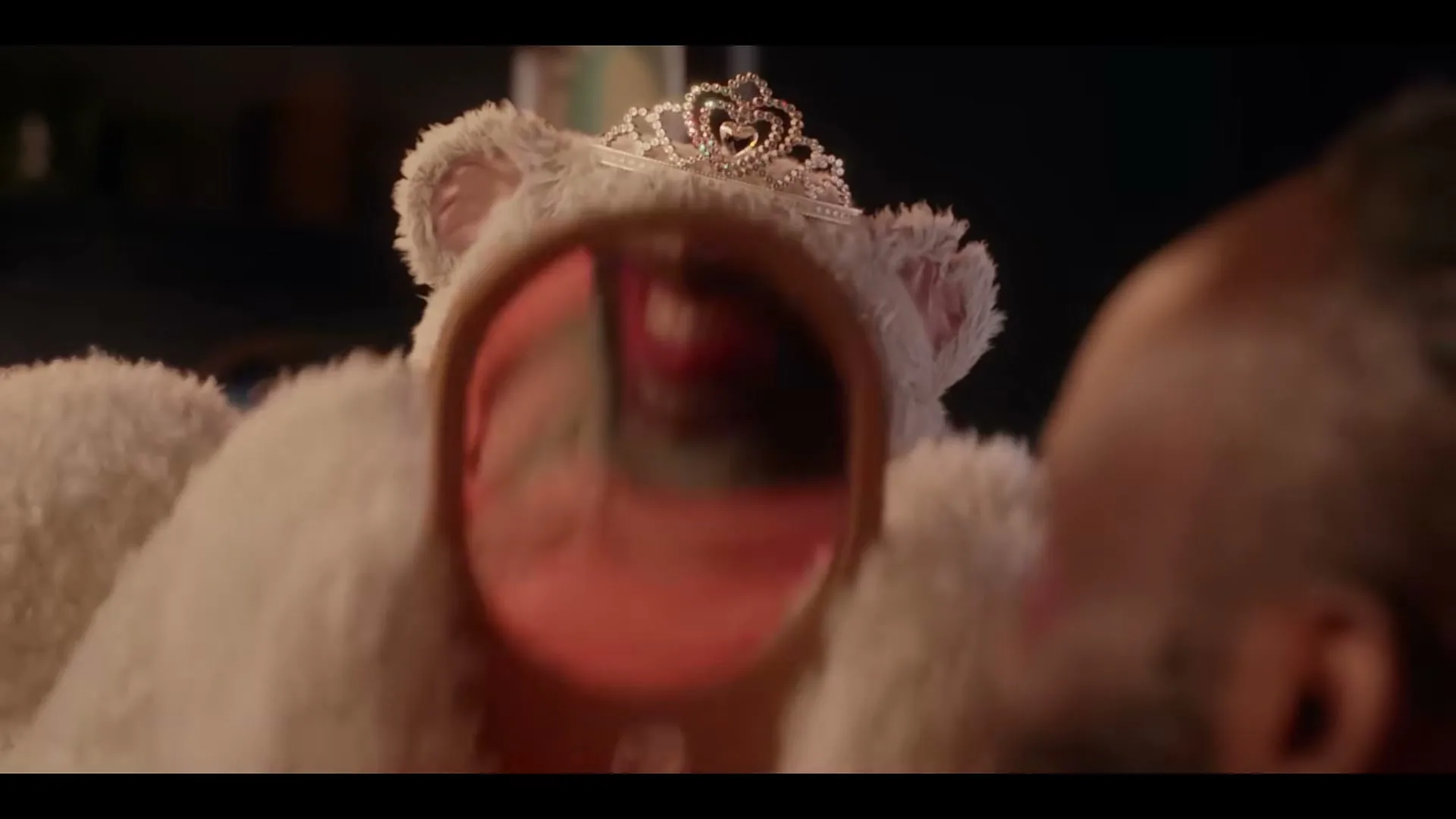 A man in a white teddy bear costume with a tiara holds a mirror to his face, which is made up with colorful makeup, then lowers the mirror to reveal the teddy bear's face, also made up, in a bedroom.