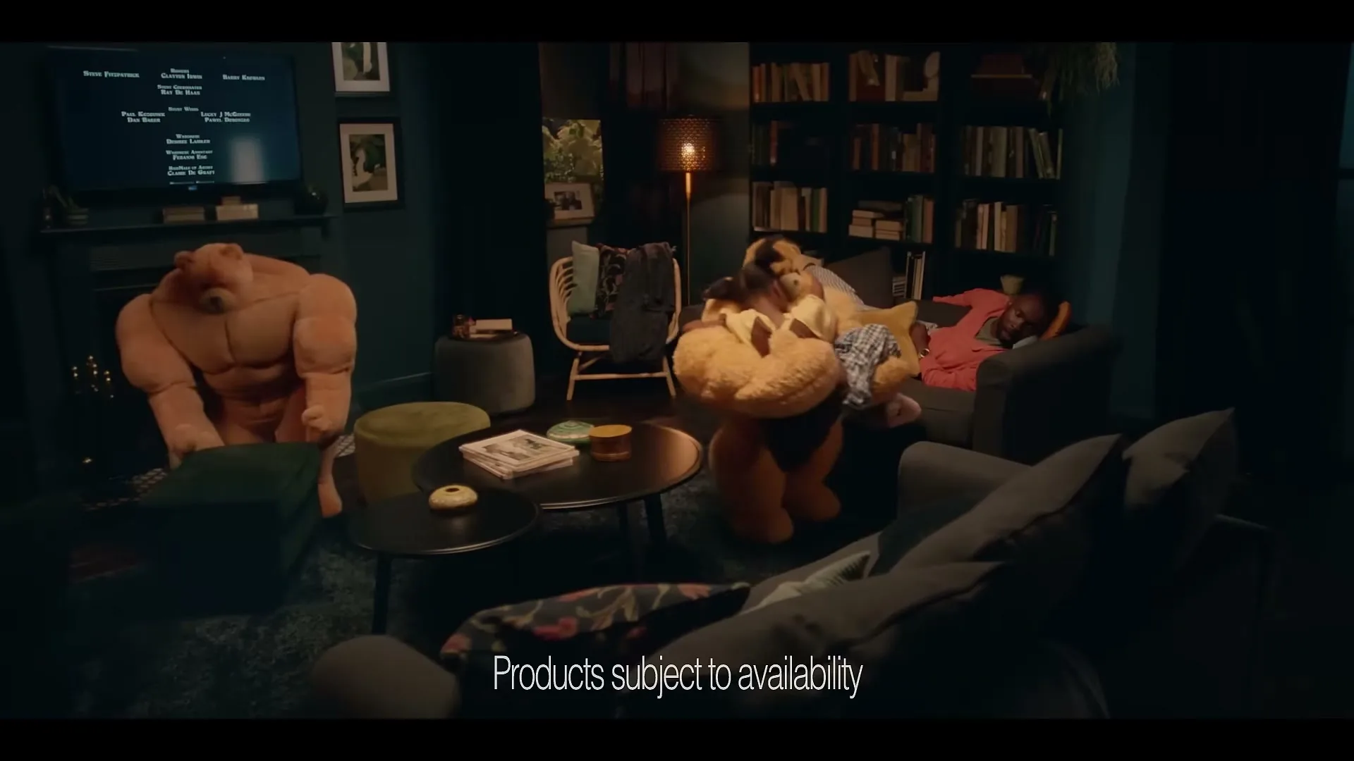 A young person sleeps on a sofa while two large teddy bears, one muscular and one carrying smaller bears, move around a cozy living room at night.