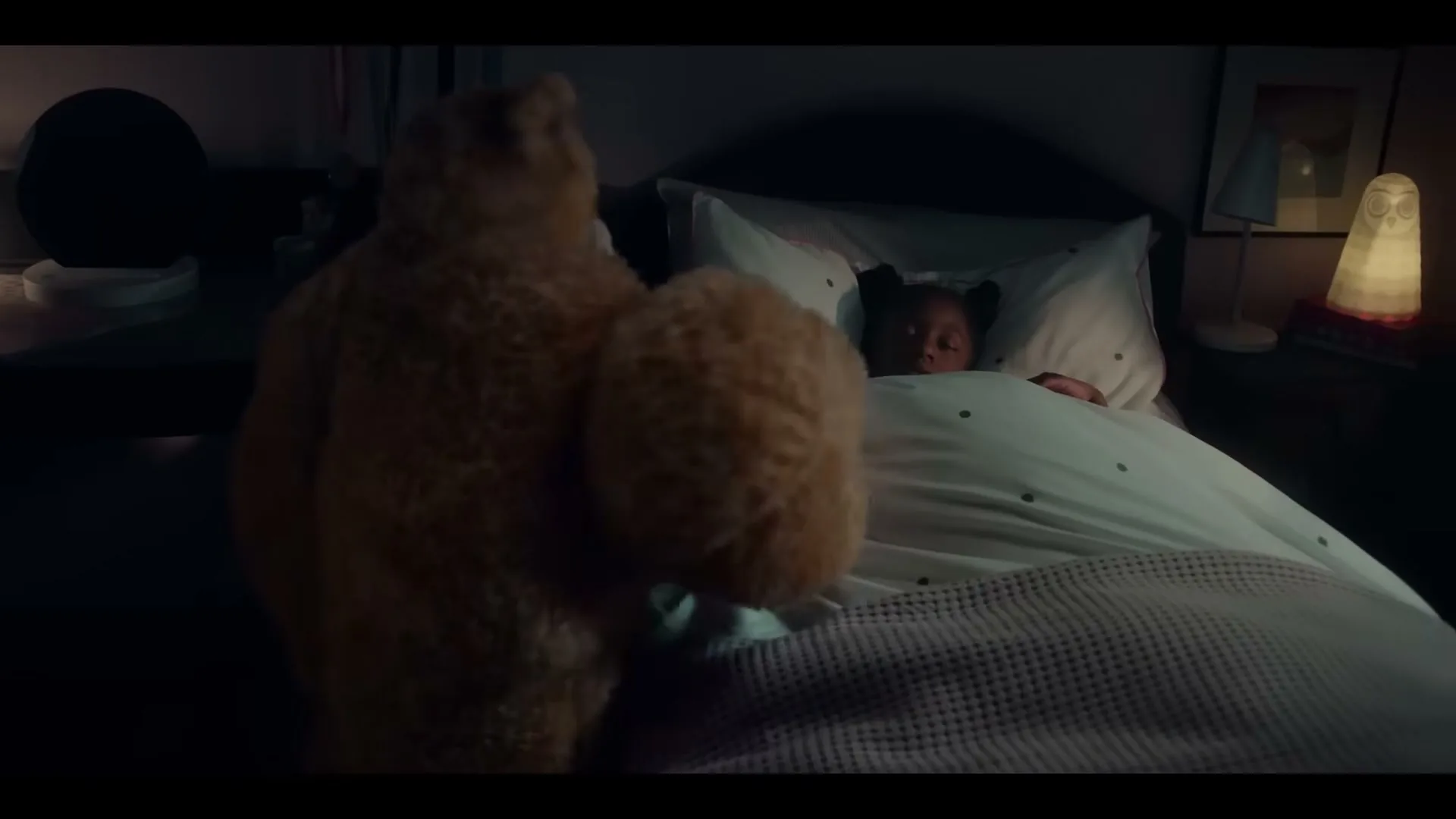 A young Black girl sleeps in her bed as a large teddy bear sits beside her in a dimly lit bedroom.