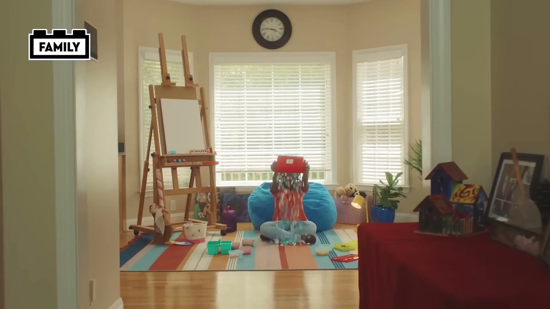 A young Black girl sits on a blue beanbag chair in a brightly lit living room, pouring a box of Lego bricks onto the floor.