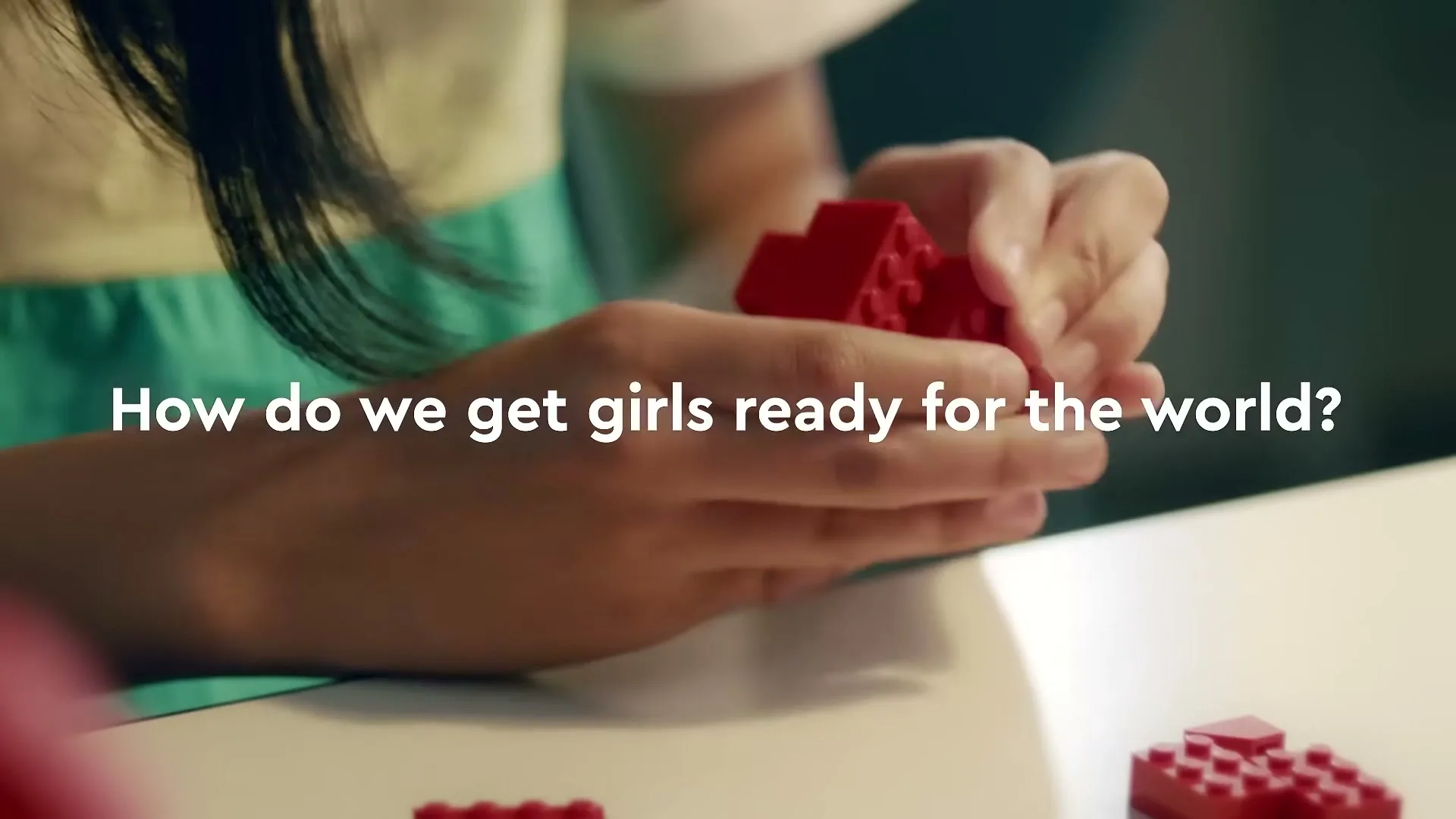 A young girl's hands are shown in a medium closeup, as she plays with red Lego bricks on a white table, with text overlay.