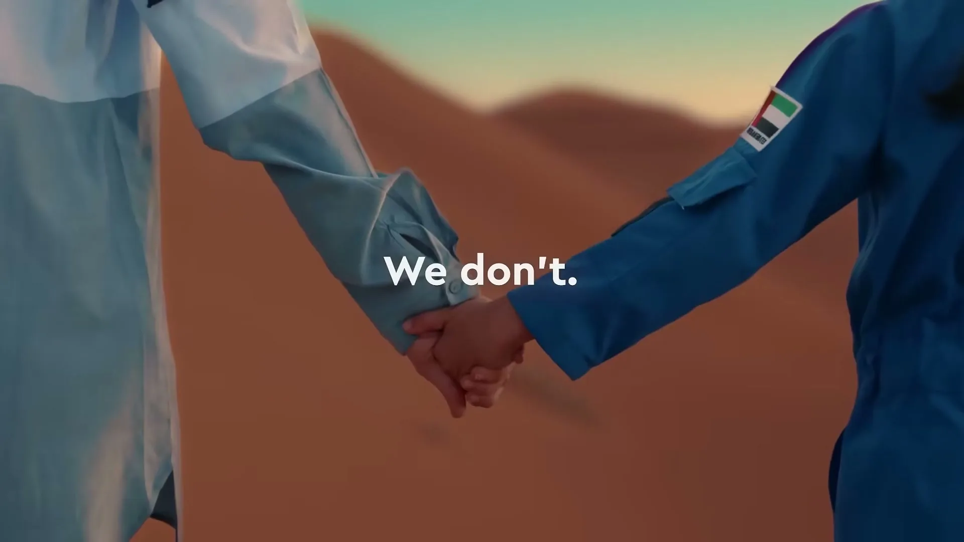 Two people, one in a white and blue robe and the other in a blue jumpsuit, hold hands in a desert landscape at golden hour.