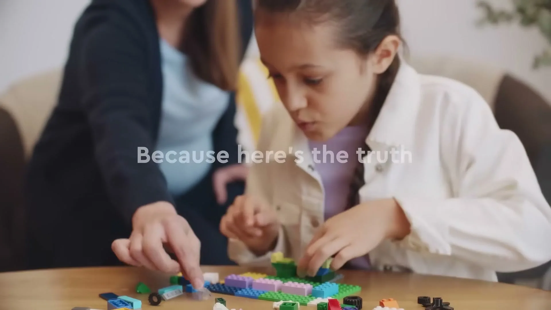 A young girl with a braid and a woman are sitting at a wooden table, building with colorful Lego bricks in an interior setting.