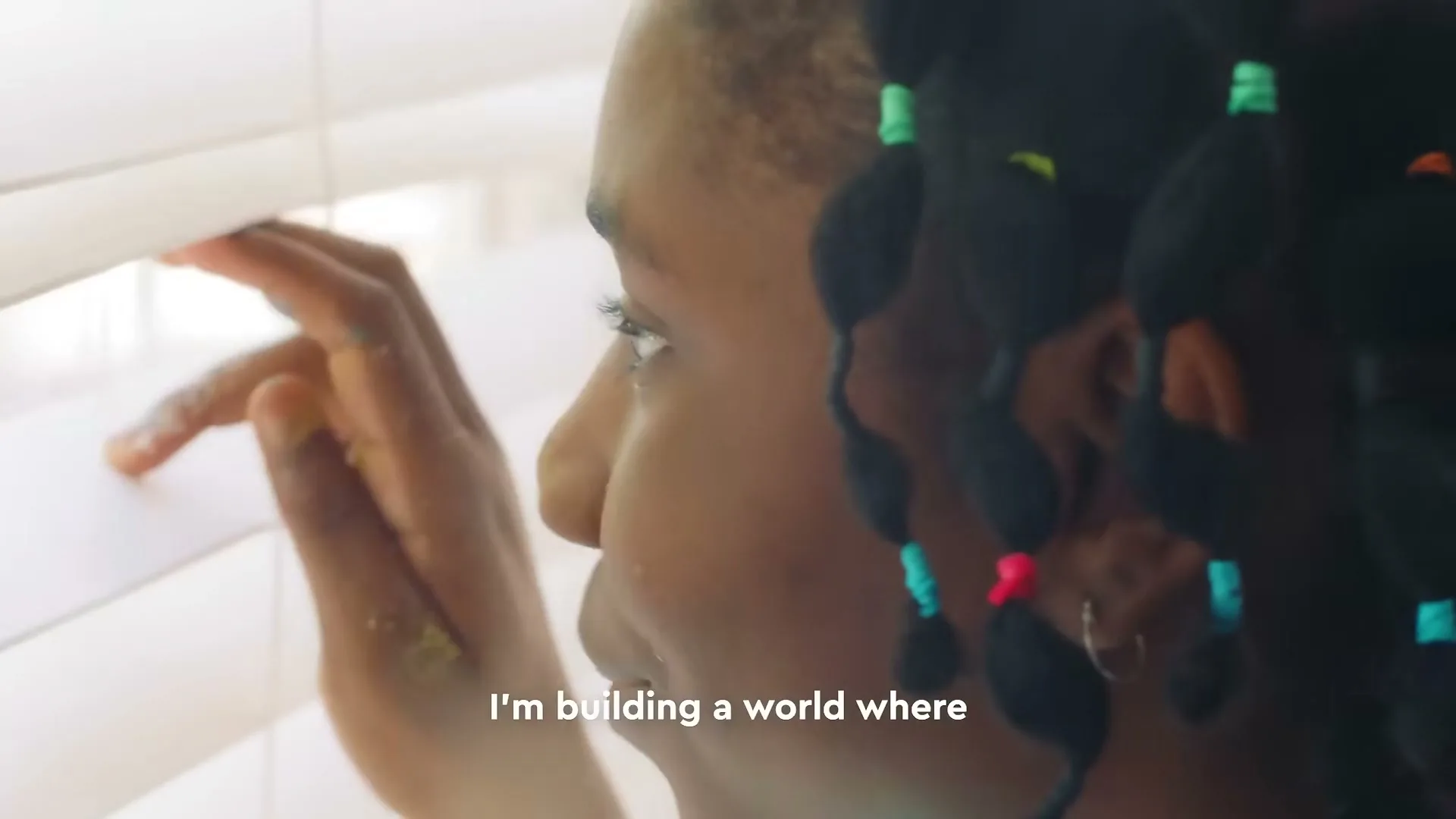 A young Black girl with braided hair looks out a window through white blinds, with yellow paint on her fingers, in a close-up shot.