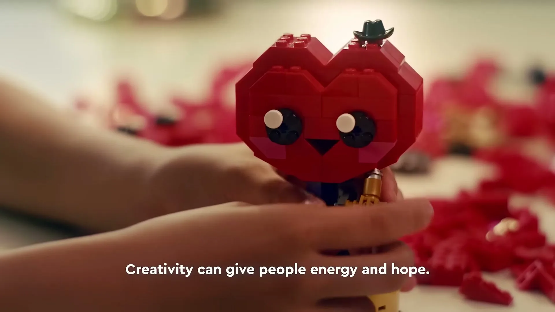 A child's hands hold a red Lego heart character with a small black hat, surrounded by scattered red Lego bricks on a light surface.