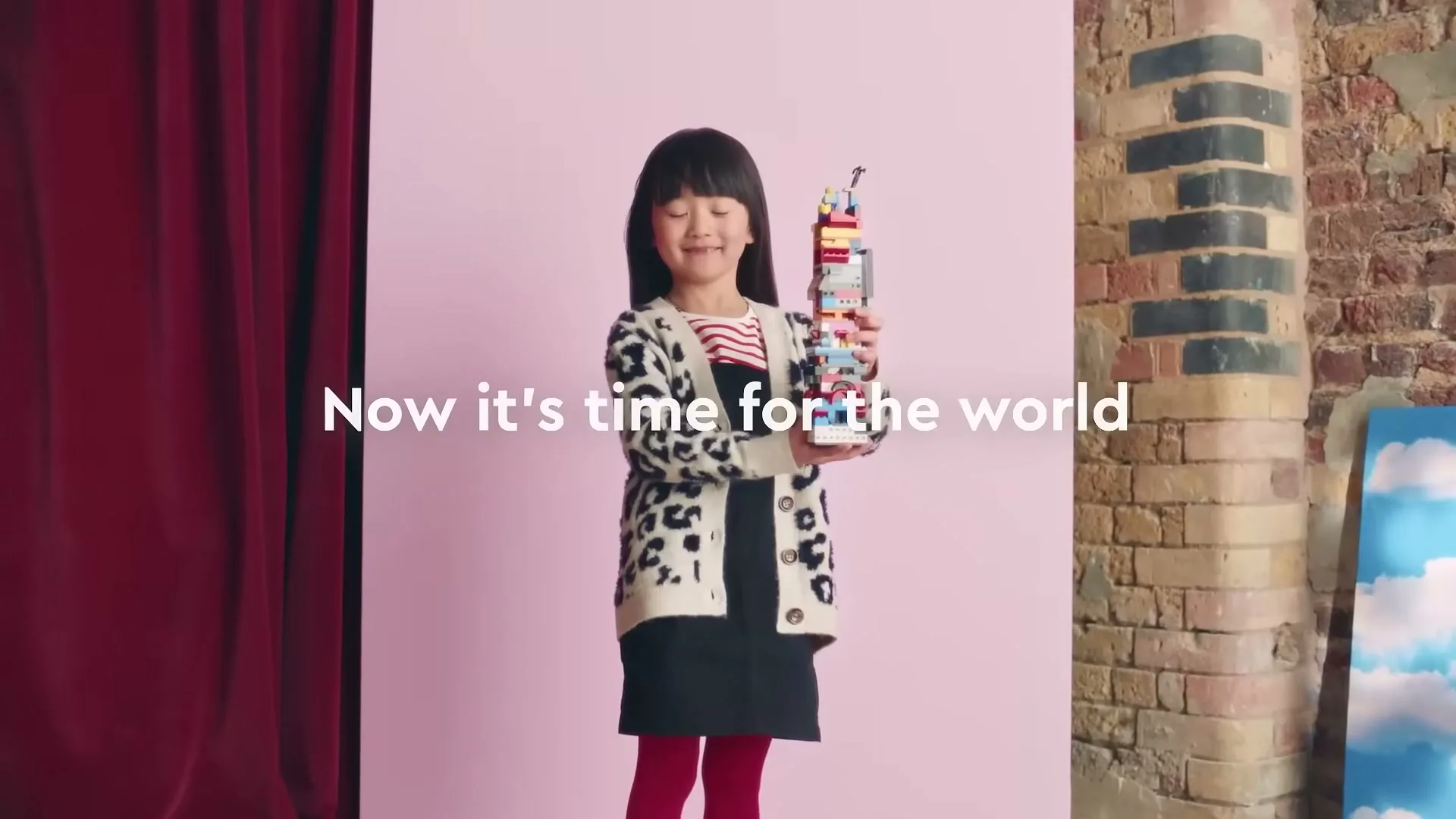 A young Asian girl in a leopard print cardigan and red tights stands smiling, holding a tall, colorful Lego creation in a studio with a pink background.
