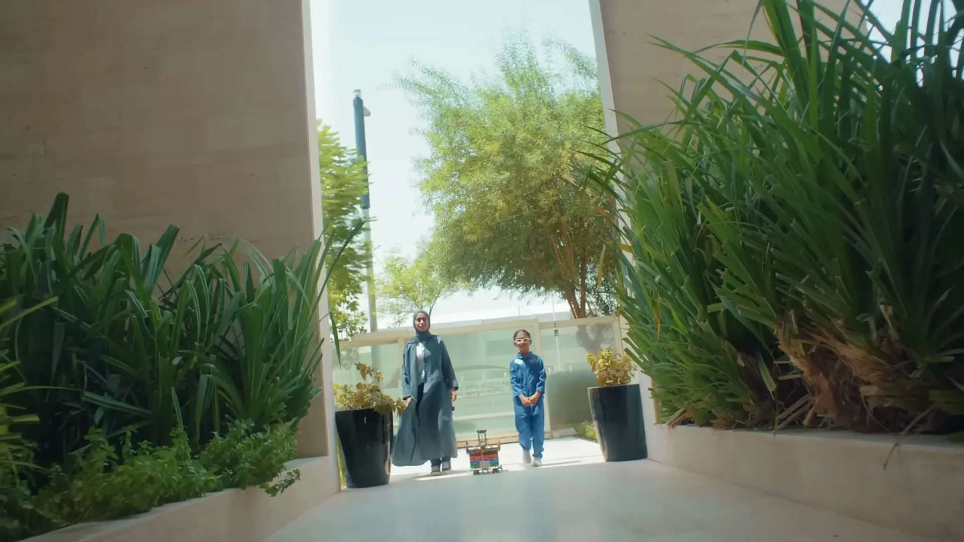 A woman in a hijab and a young girl in a blue jumpsuit walk towards the camera, with a Lego creation between them, in an outdoor walkway with lush green plants.