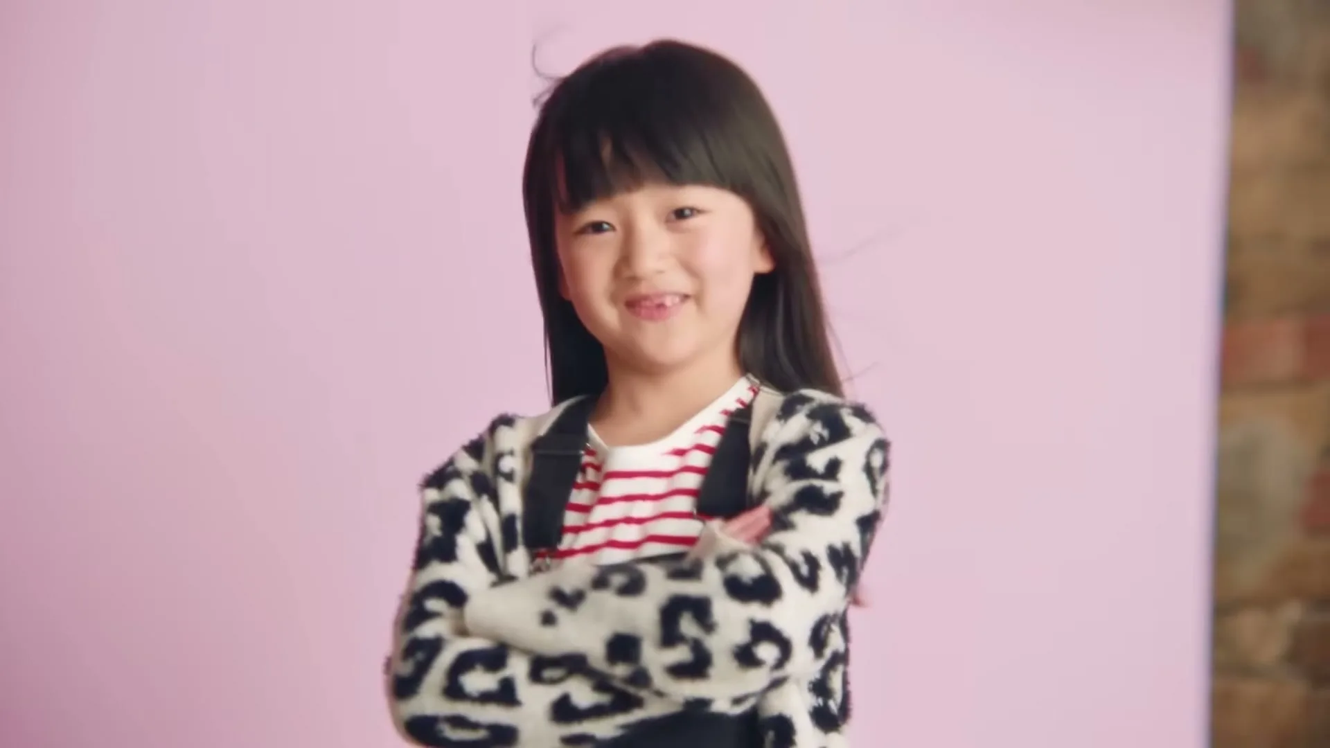 A young Asian girl with long dark hair, wearing a leopard print cardigan and striped shirt, stands with arms crossed and smiles at the camera against a pink background.