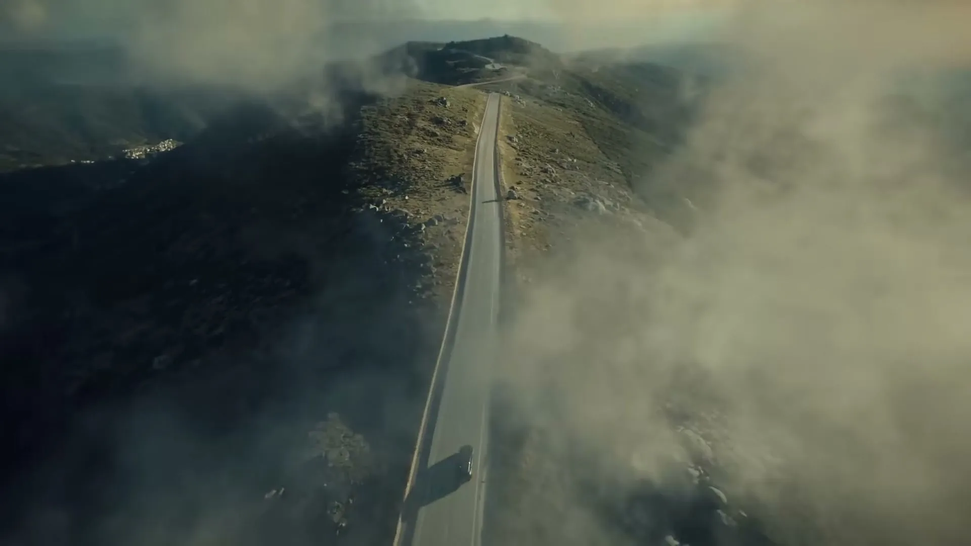 An aerial view shows a black Lexus car driving on a long, winding road through a mountainous landscape, partially obscured by clouds.