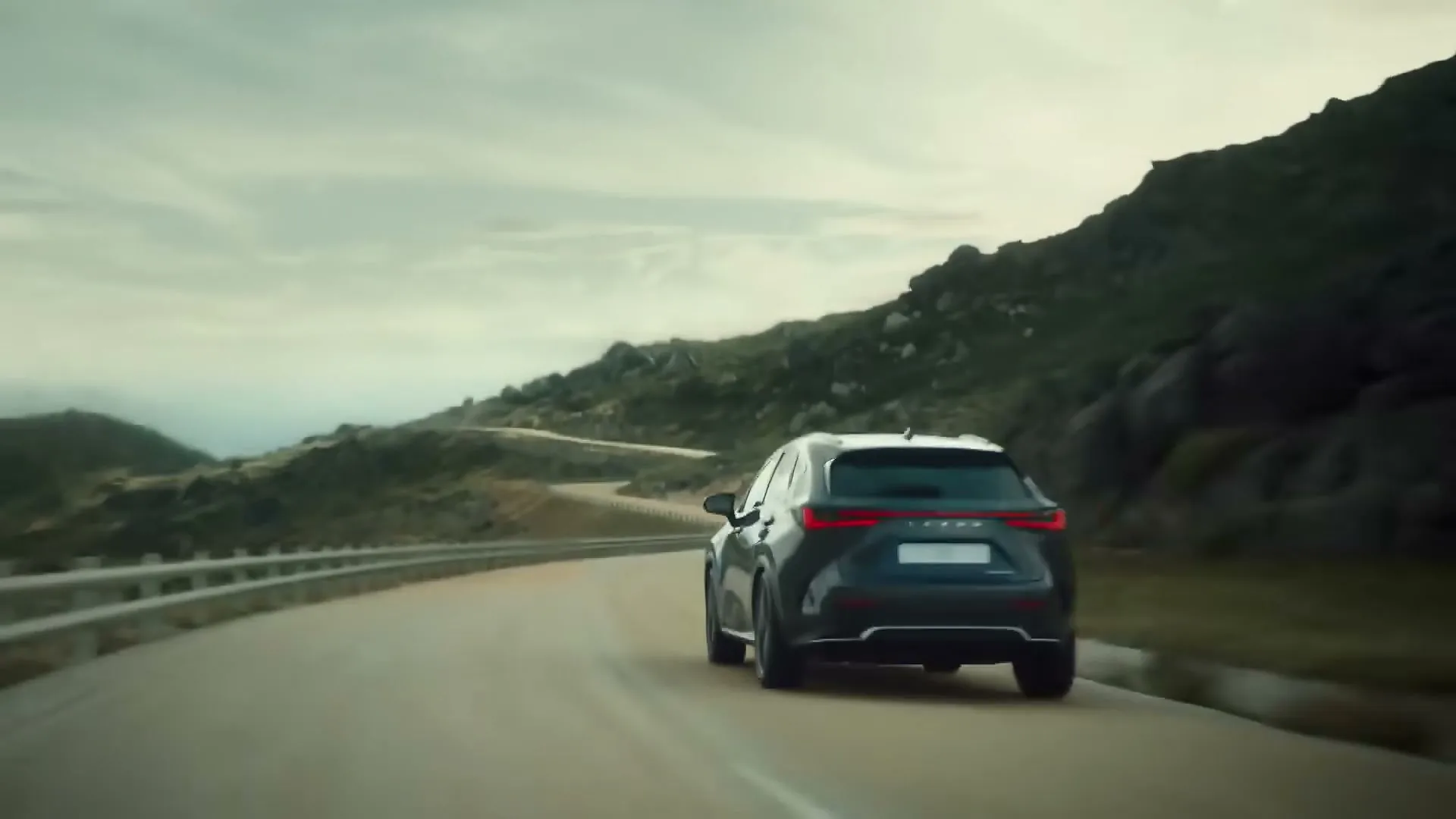 A dark grey Lexus SUV drives away from the camera on a winding mountain road under a cloudy sky.