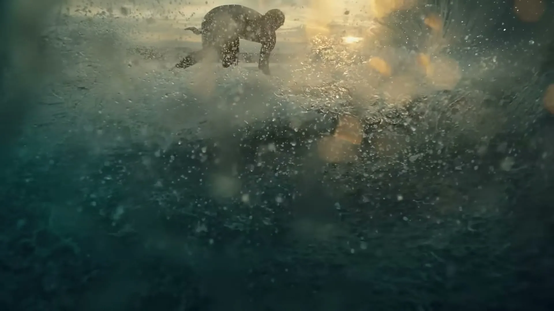 A surfer in a wetsuit rides a wave at sunset, viewed from underwater with water splashing in the foreground.