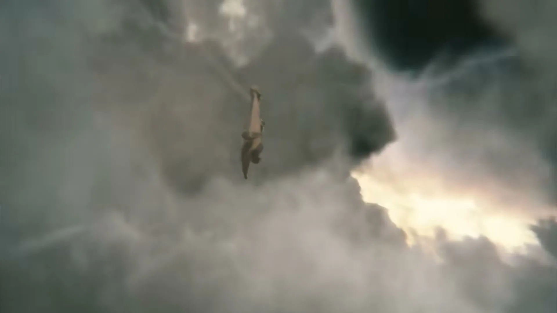 A person in a wingsuit is seen falling through a cloudy sky, with sunlight breaking through the clouds in the background.