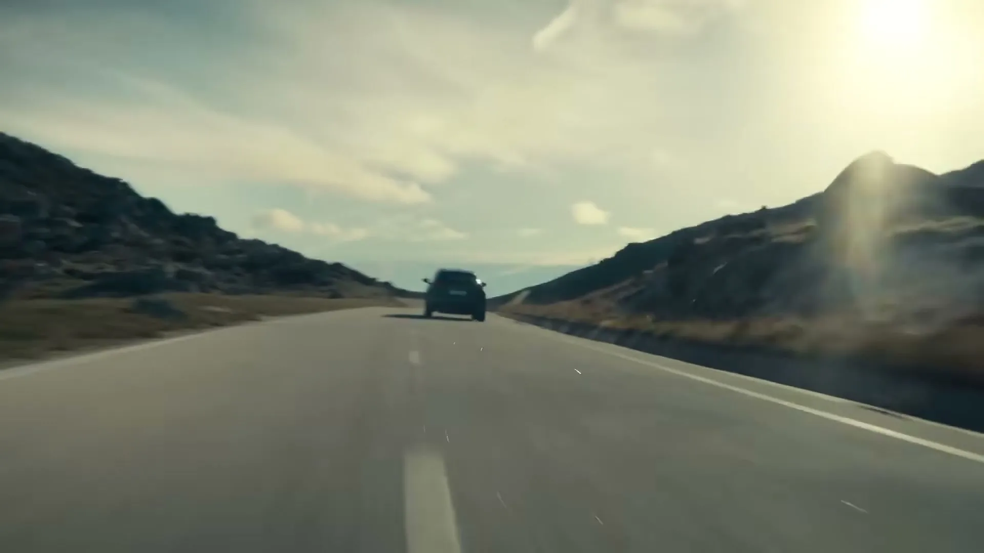 A dark Lexus SUV drives away from the camera on a winding mountain road under a bright, sunny sky.