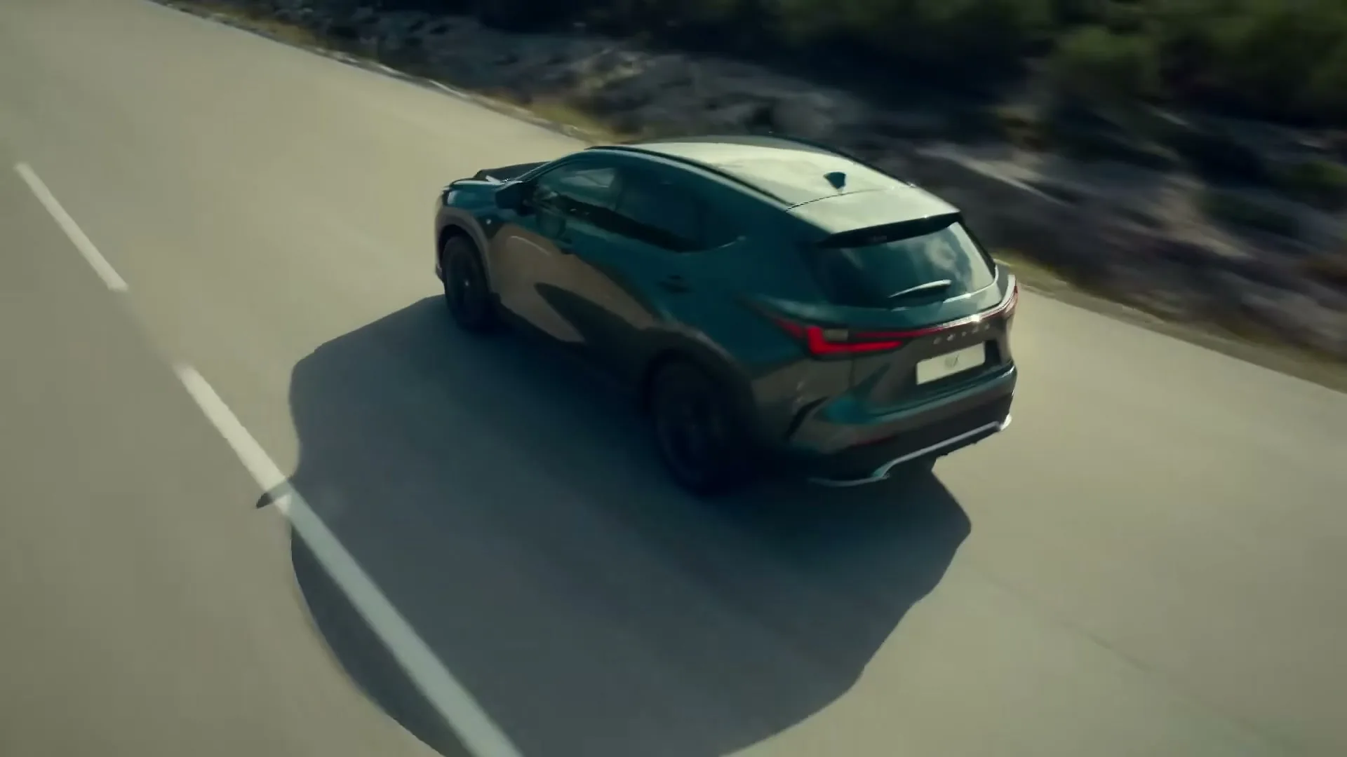 A dark green Lexus SUV drives on a winding road with a long shadow, captured from an overhead perspective.