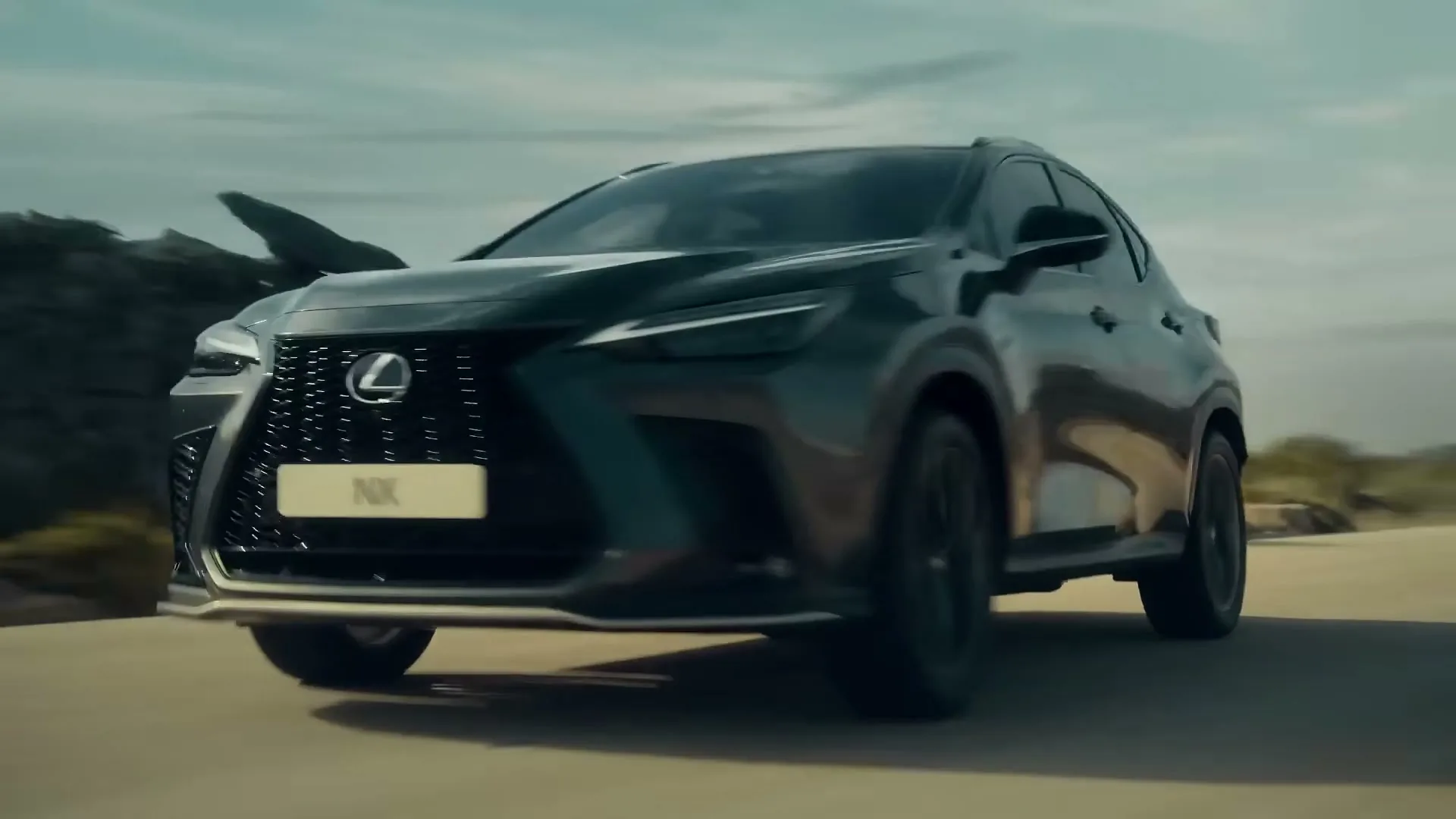A dark grey Lexus NX SUV drives quickly on a paved road under a cloudy sky, with blurred background elements suggesting speed.