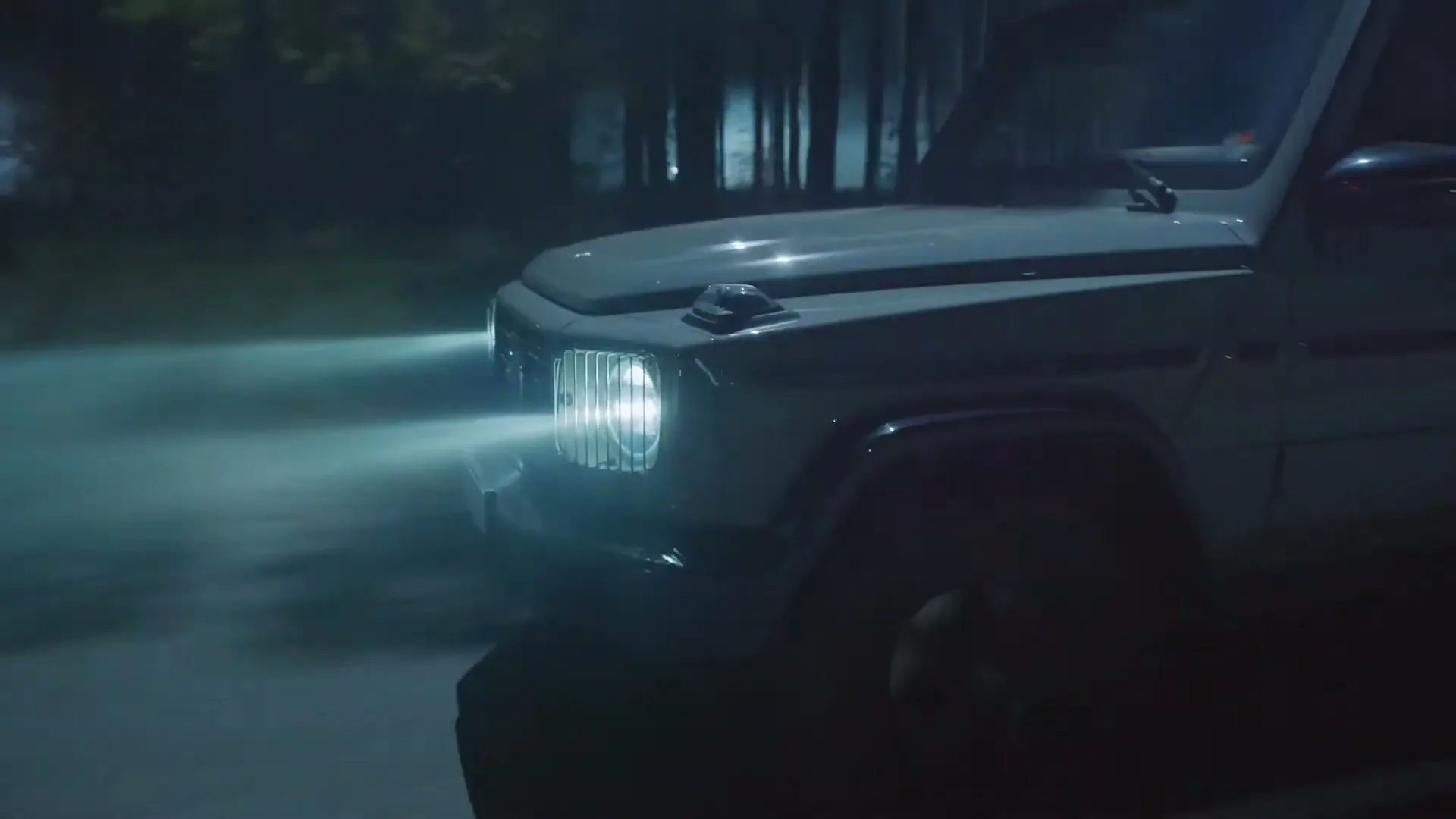 A dark Mercedes G-Wagen drives through a forest at night, its headlights illuminating the path.