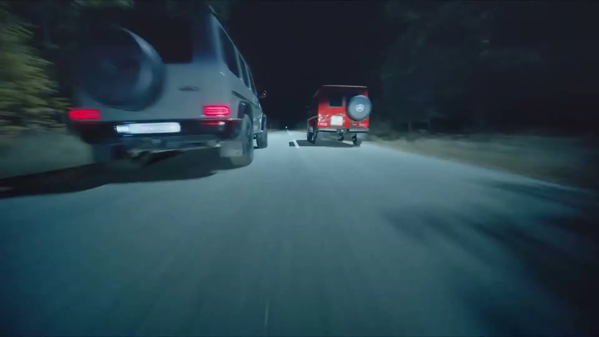 Two Mercedes G-Wagens, one grey and one red, drive at night on a dark, tree-lined road.