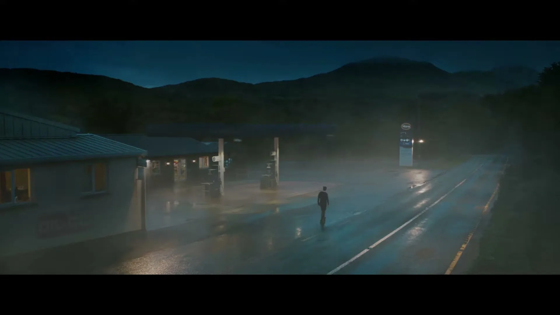 A lone man stands in the middle of a wet, misty road outside a gas station at night, with mountains in the background.