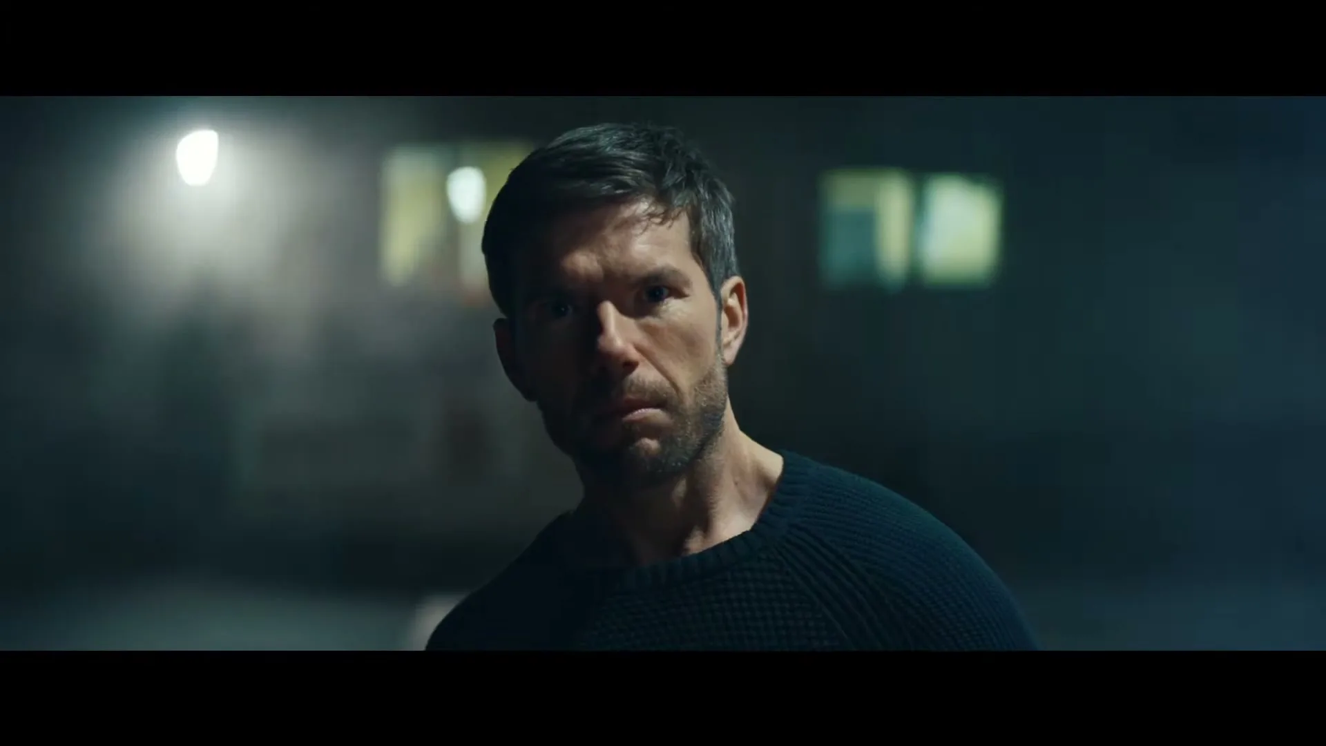A man with dark hair and a beard looks intently at the camera in a dimly lit, smoky interior space.
