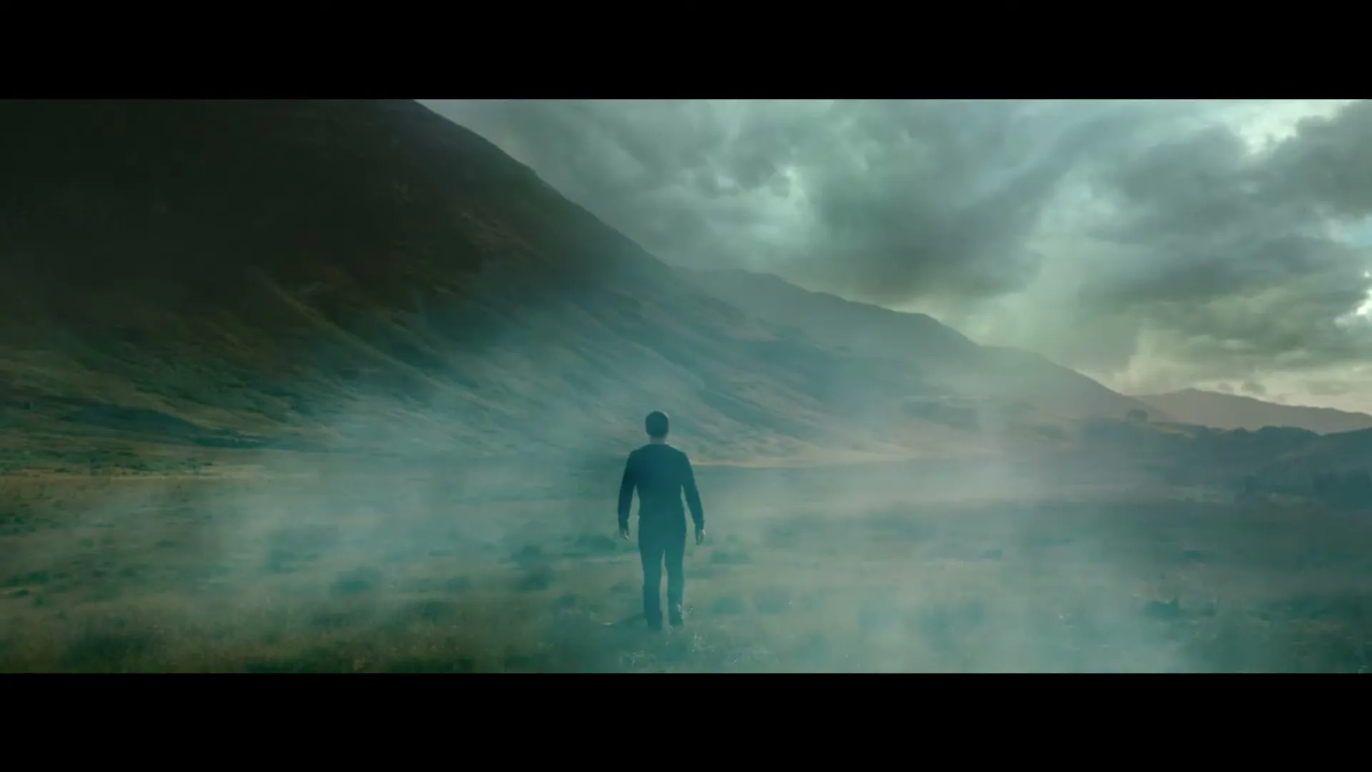 A lone man stands in a vast, misty valley surrounded by mountains under a dramatic sky, looking into the distance.