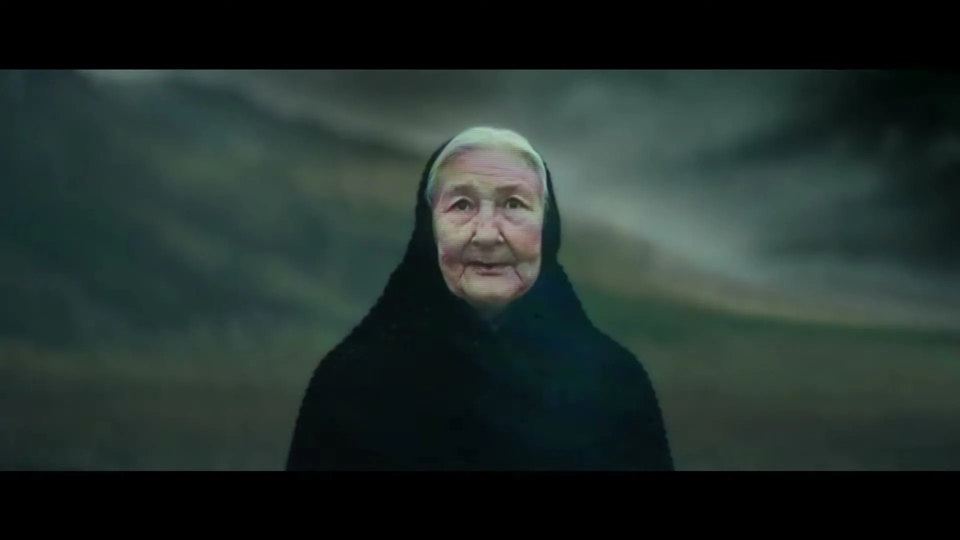 An elderly woman with white hair and a black shawl looks directly at the camera, standing against a blurred, dark green landscape.