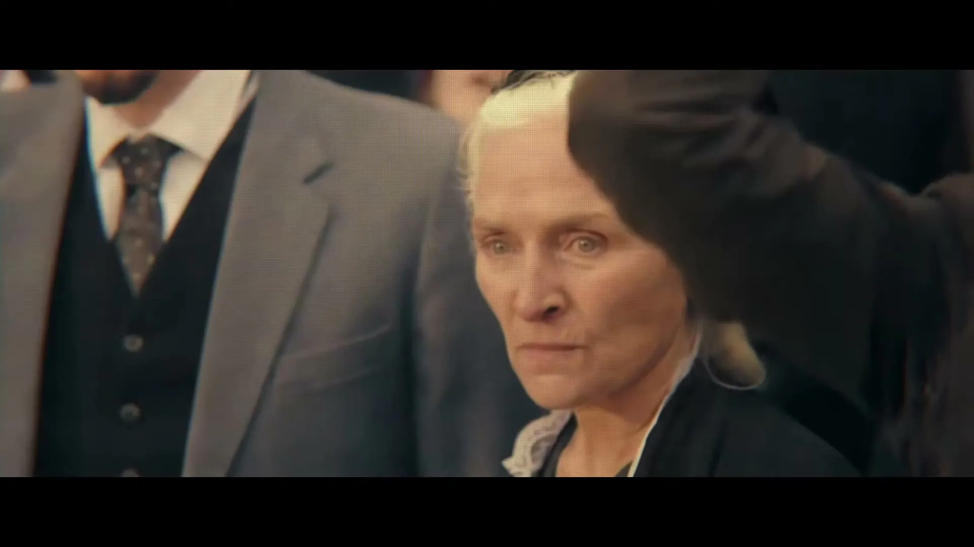 An older woman with white hair and a black dress looks intently forward, surrounded by people in suits, in an interior setting.