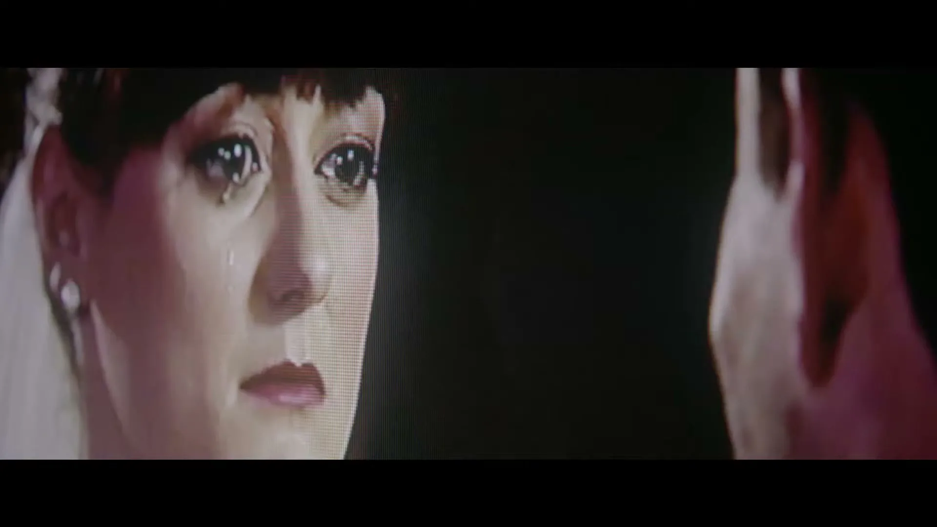 A woman with dark hair and a white veil cries, a tear rolling down her cheek, as she looks at a blurry figure in a dark room.